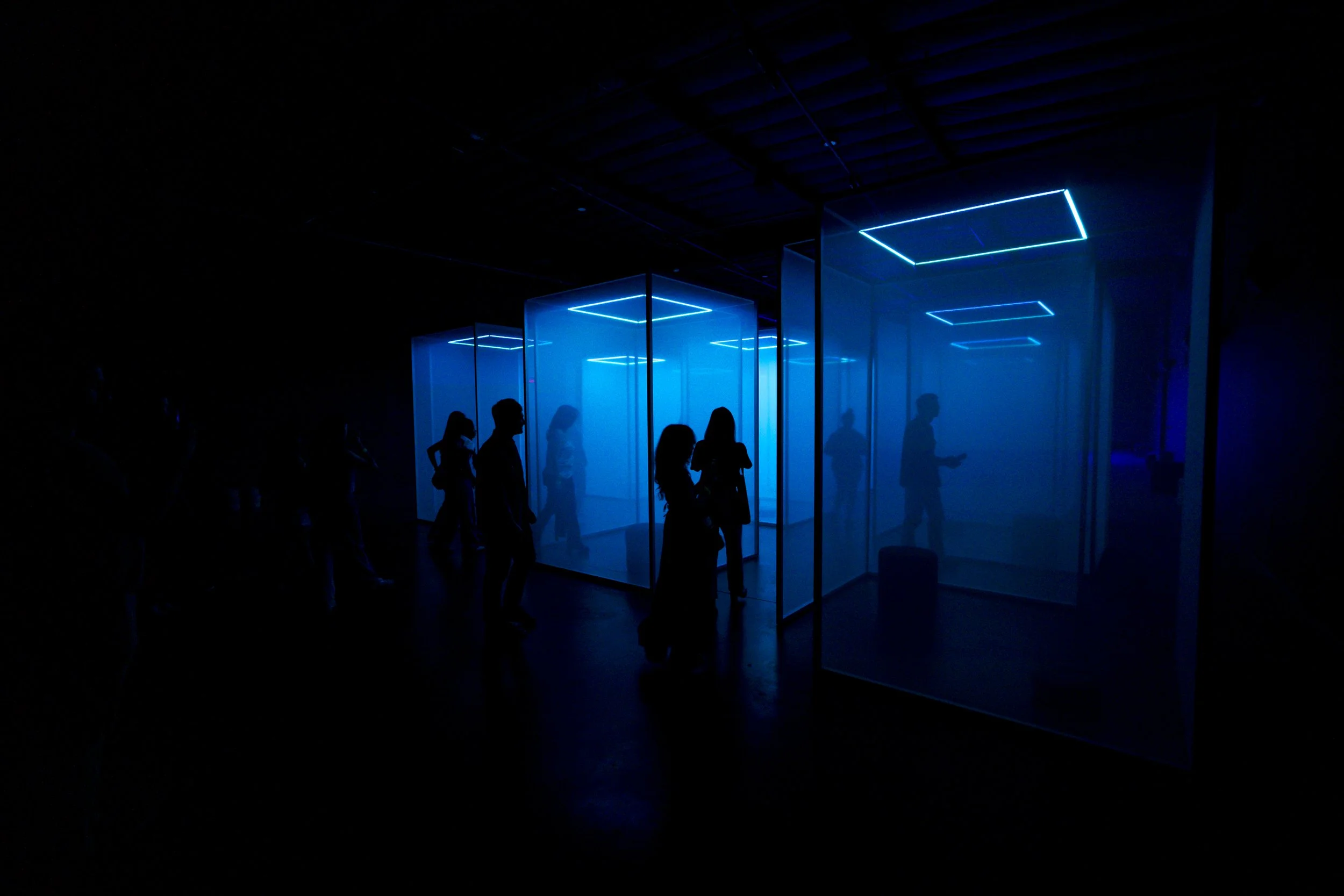 People in silhouette walk past illuminated glass exhibition booths with blue neon lights in a dark space.