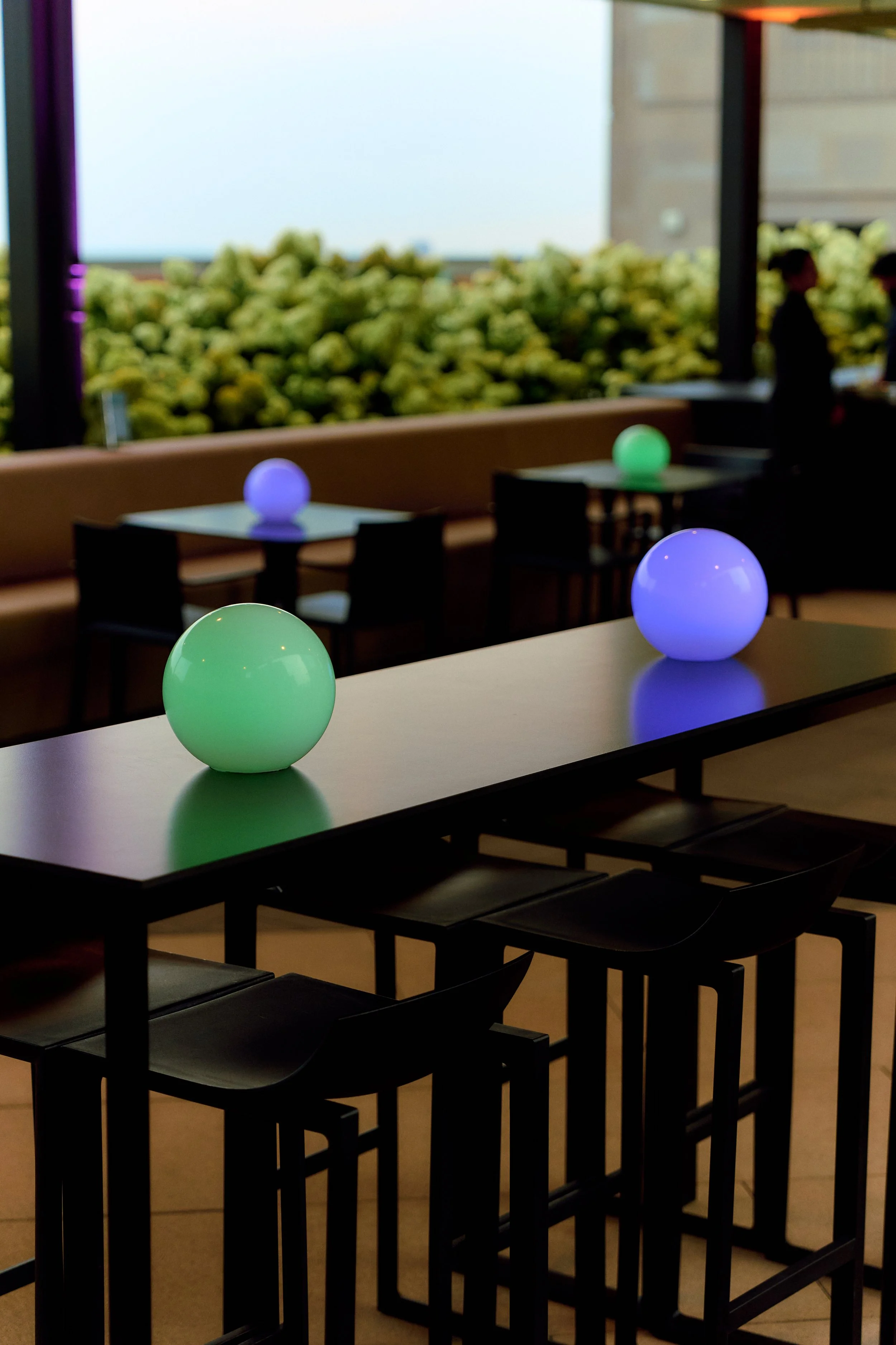 Modern art installation featuring glowing green and purple spheres on black tables in a dimly lit dining area with large window and greenery outside.