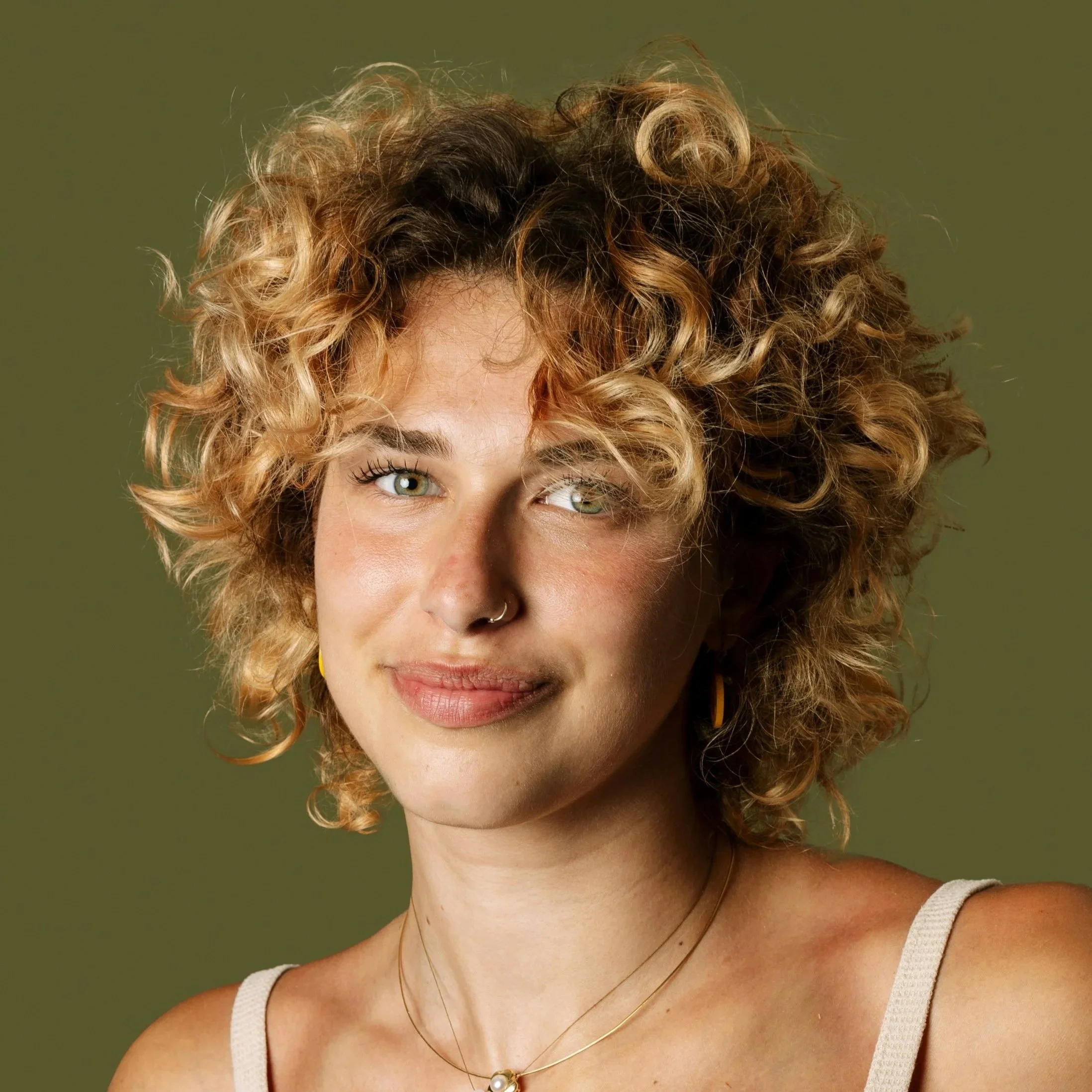 Close-up portrait of a woman with curly blonde hair, green eyes, wearing a beige tank top and layered necklaces, against a plain olive green background.