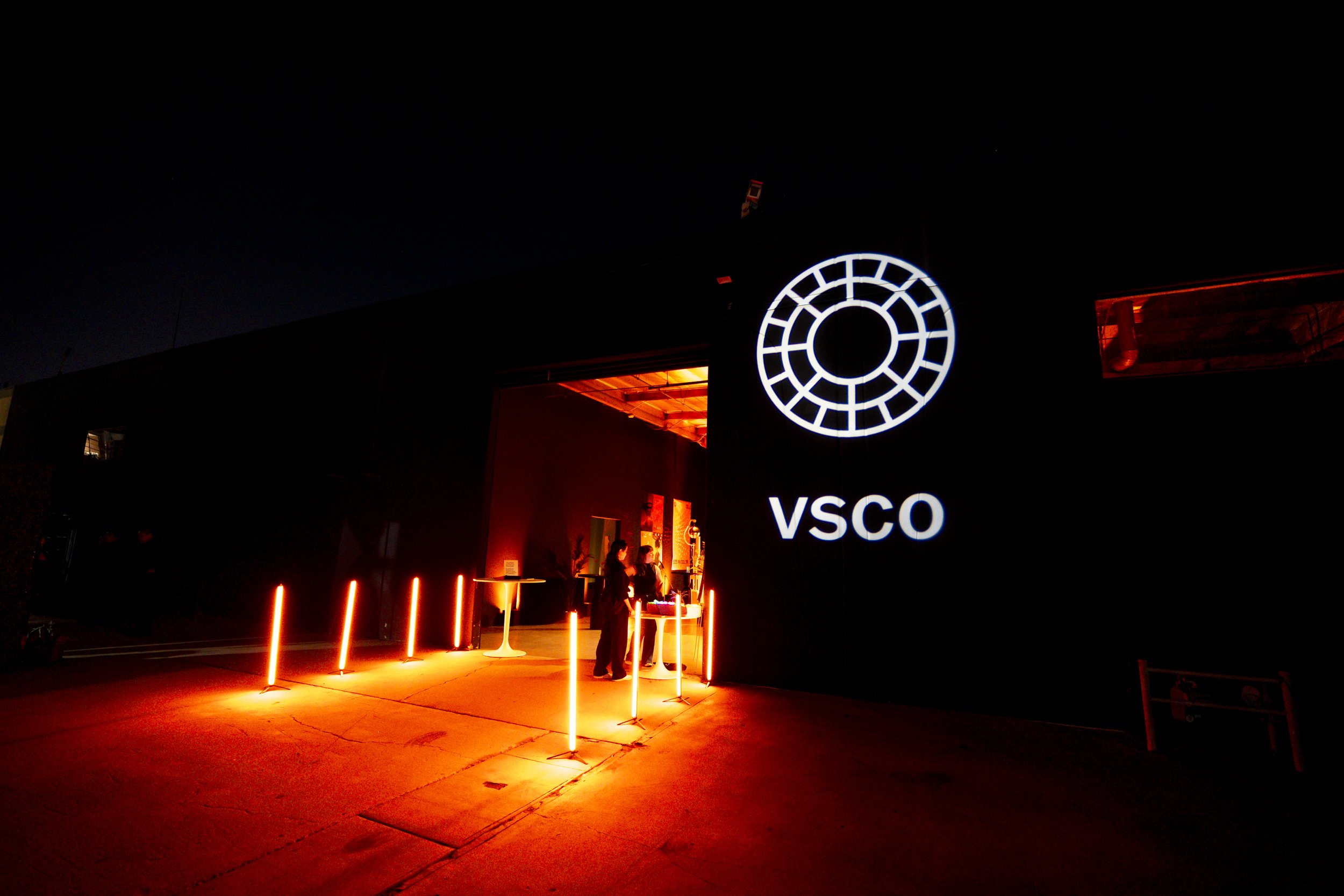 Nighttime entrance to an event space with illuminated vertical red lights and a large sign featuring a white circular logo and the text 'VSCO'.