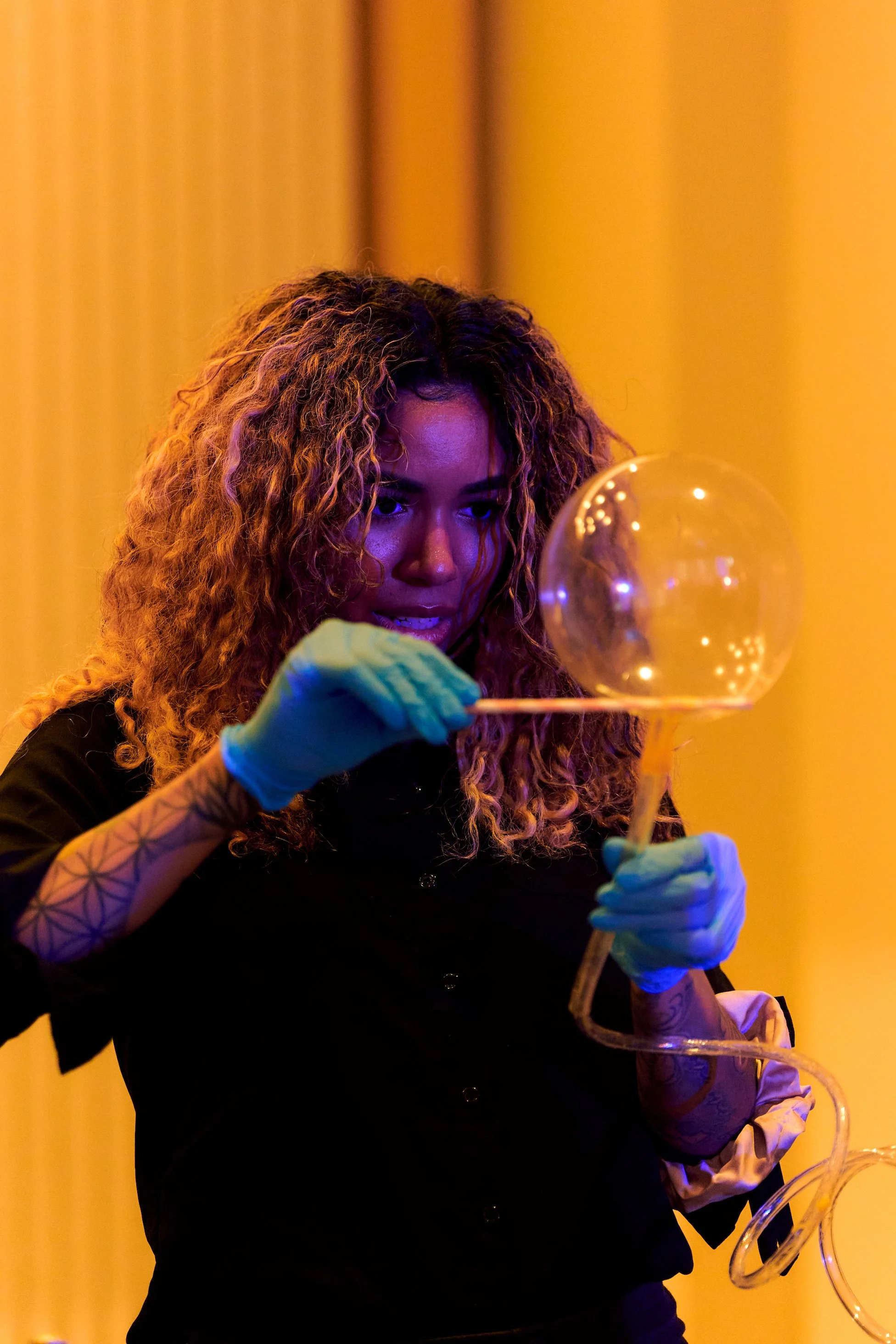 A woman with curly hair and blue gloves is shaping a large glass bubble with a tool.
