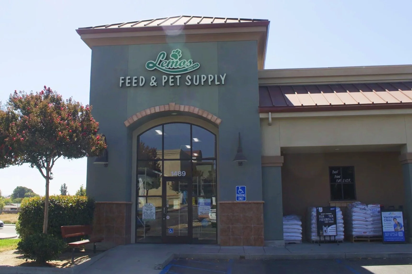 Exterior view of Paso Robles Lemos Feed & Pet Supply store with large glass entrance doors, a handicap parking space, and a tree on the left. The store sign is above the entrance. The parking lot is visible with marked parking spots.