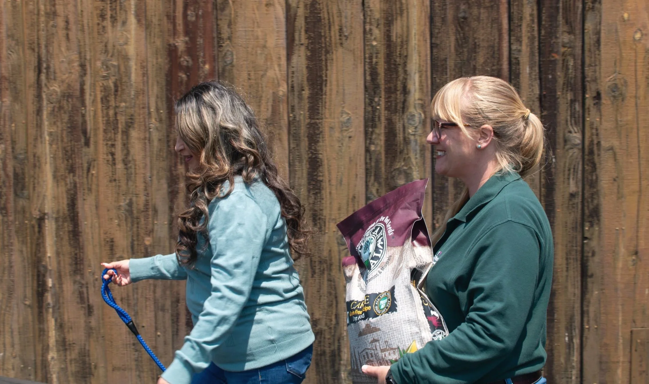 Two women walking outdoors in front of a wooden fence, one holding a bag of pet food, both smiling and talking.
