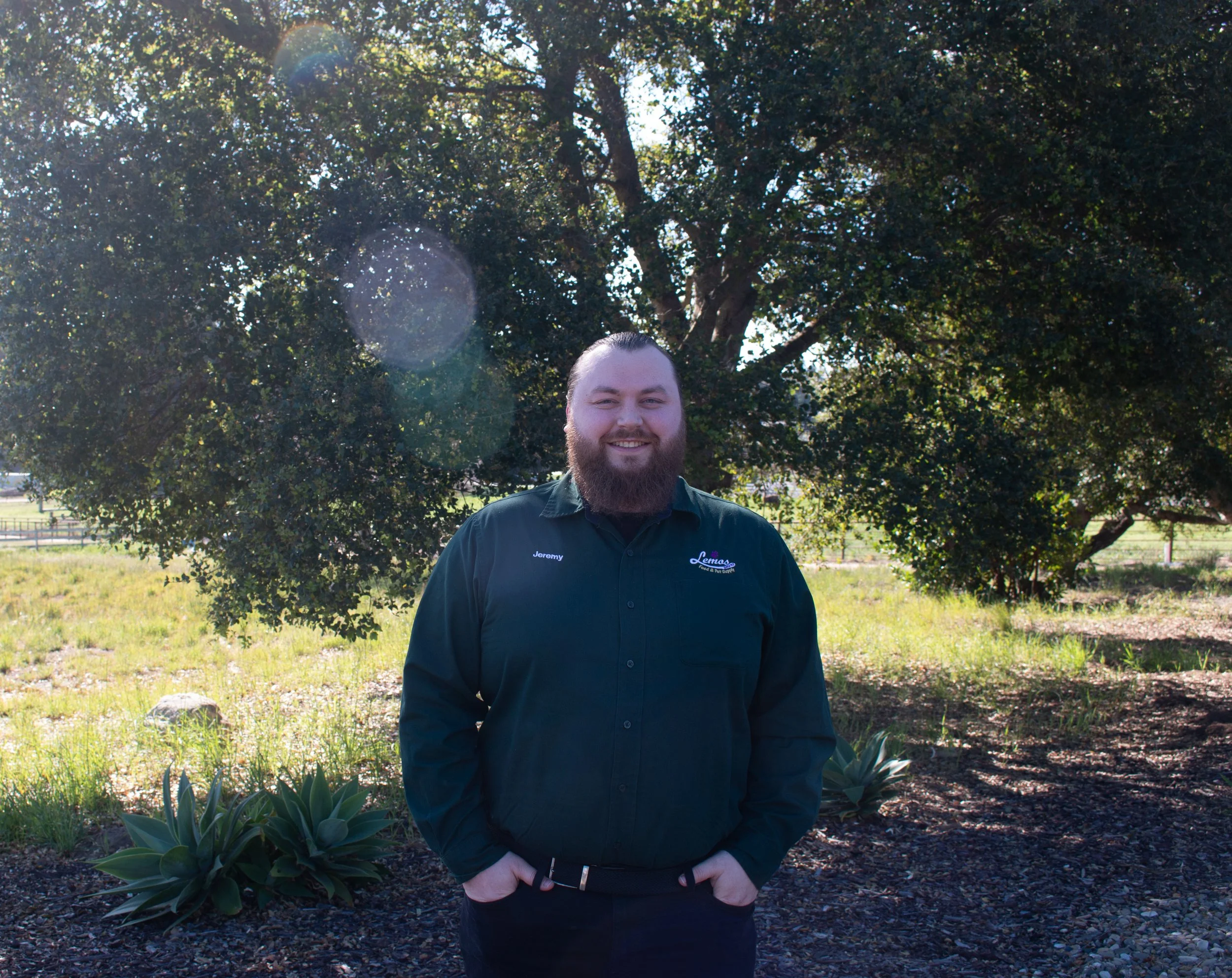 A man with a beard and long hair, wearing a black shirt with the name Jeremy embroidered on it, standing outdoors in front of a large tree with lush foliage, smiling at the camera.