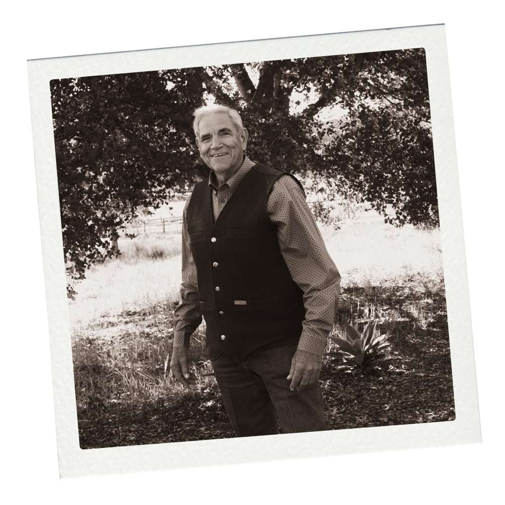 Black and white photograph of an older man standing outdoors under a tree, smiling. He is wearing a patterned shirt and a dark vest, with a background of grass and plants.