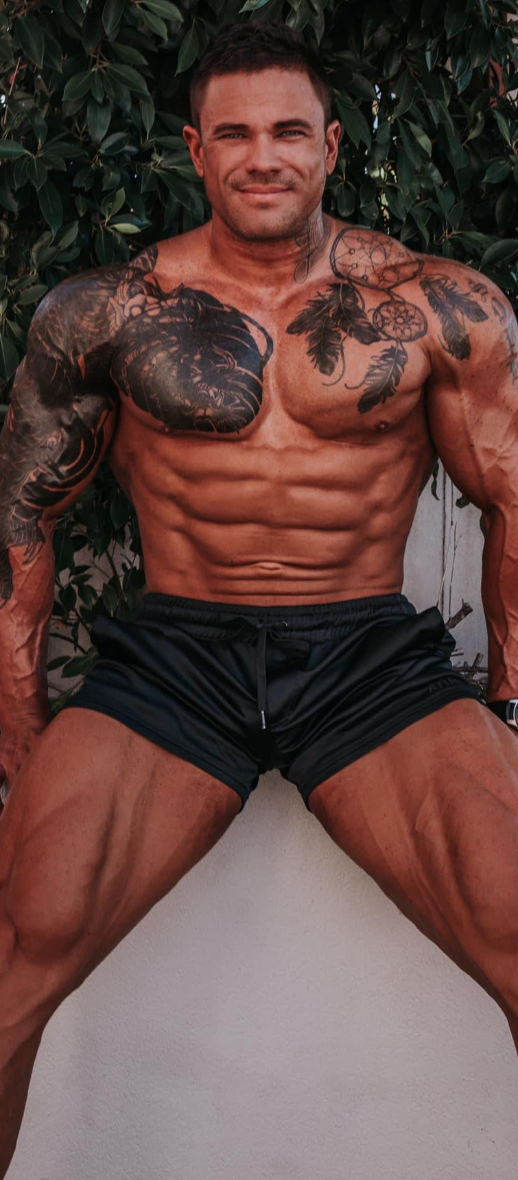 Liam a tattooed man posing outdoors with foliage background.