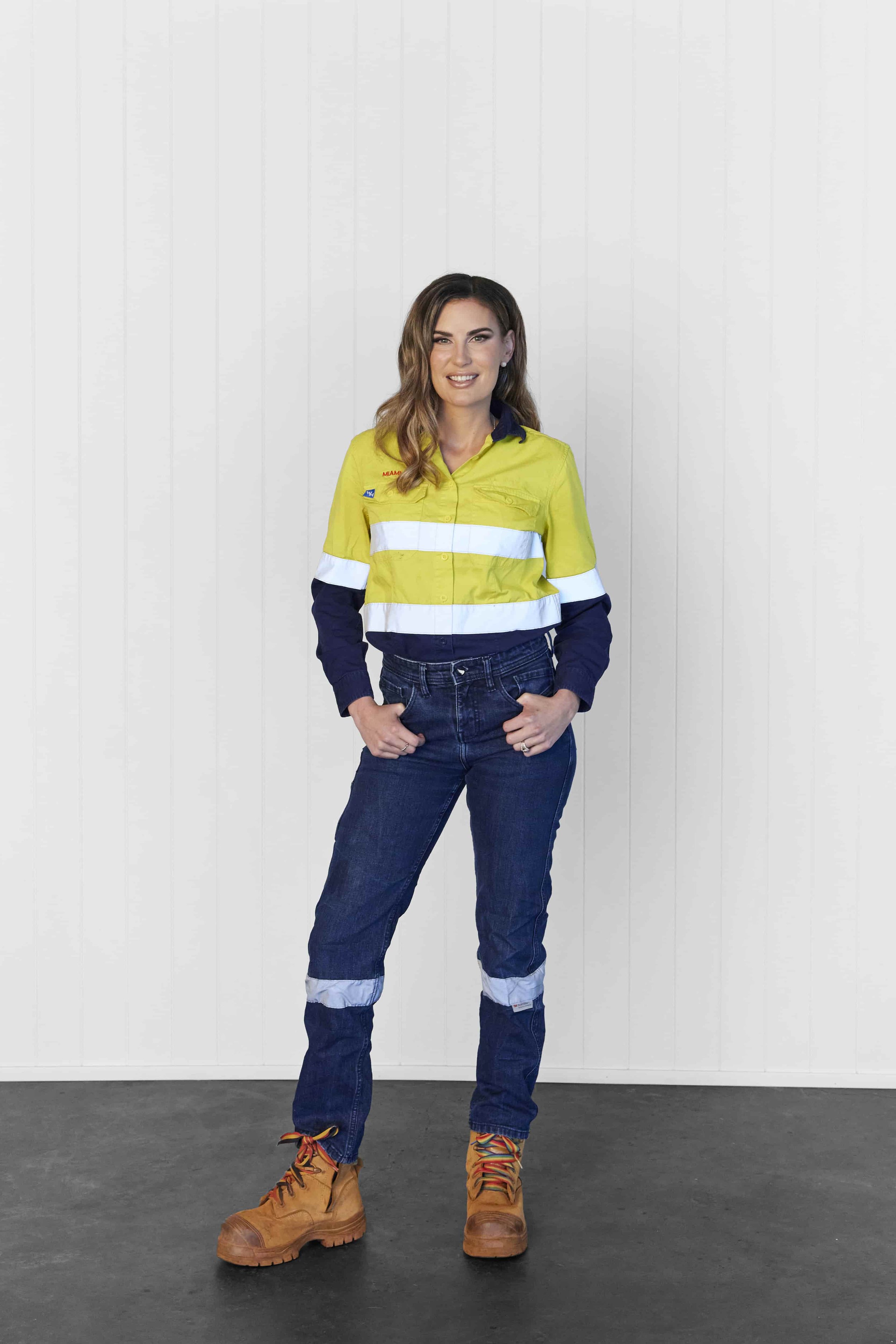 Woman wearing a high-visibility yellow and navy work shirt, jeans with reflective patches, tan work boots, standing with hands in pockets against a white paneled wall.