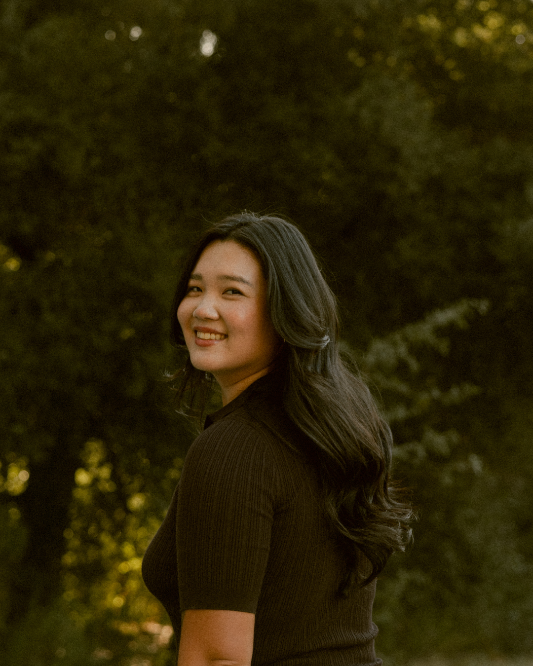 A smiling woman with long dark hair standing outdoors with trees in the background.