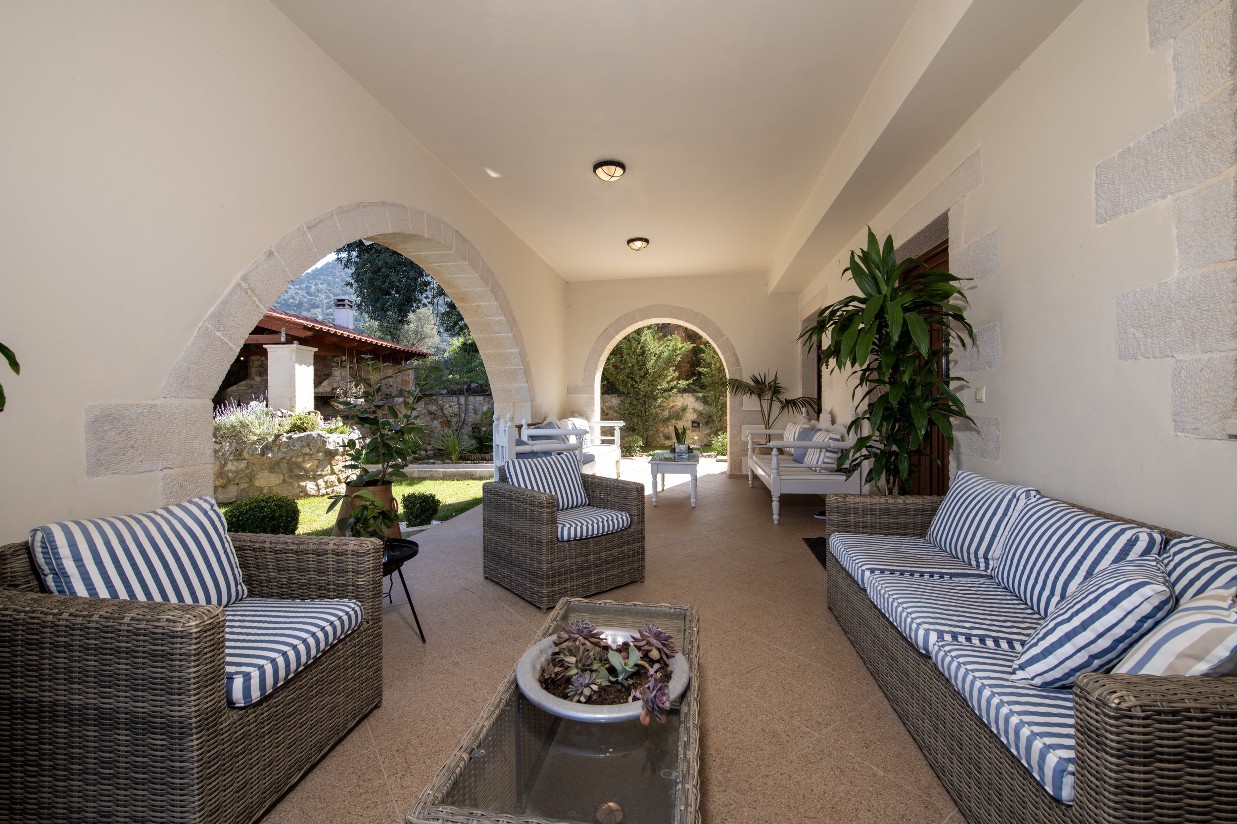 Covered patio with wicker furniture, striped cushions, arched openings, and potted plants.