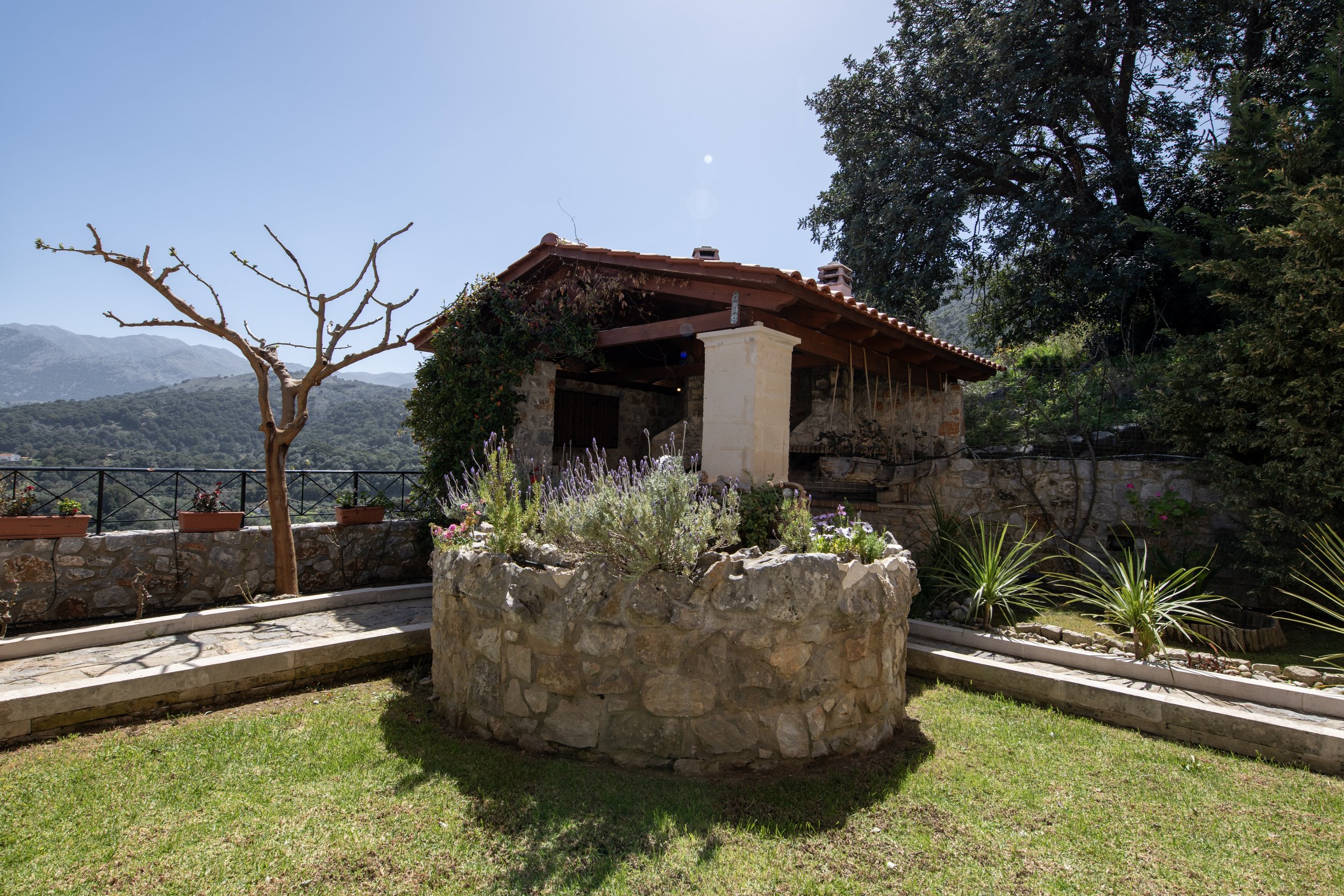 Stone patio with a circular herb garden, a tree with no leaves, a rustic wooden building with a red roof, and a scenic mountain view in the background.
