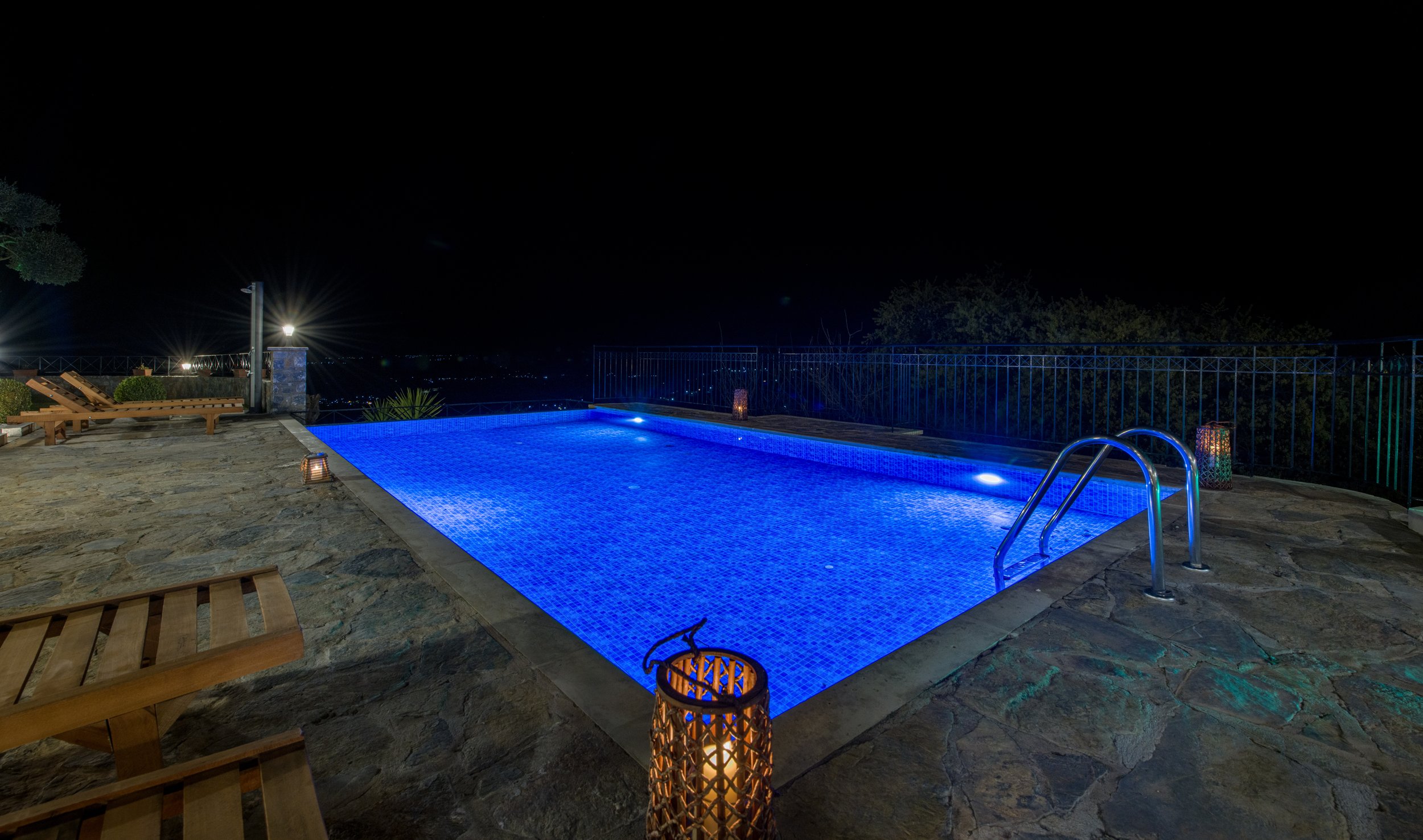 Illuminated swimming pool at night with deck chairs and lanterns on a stone patio.
