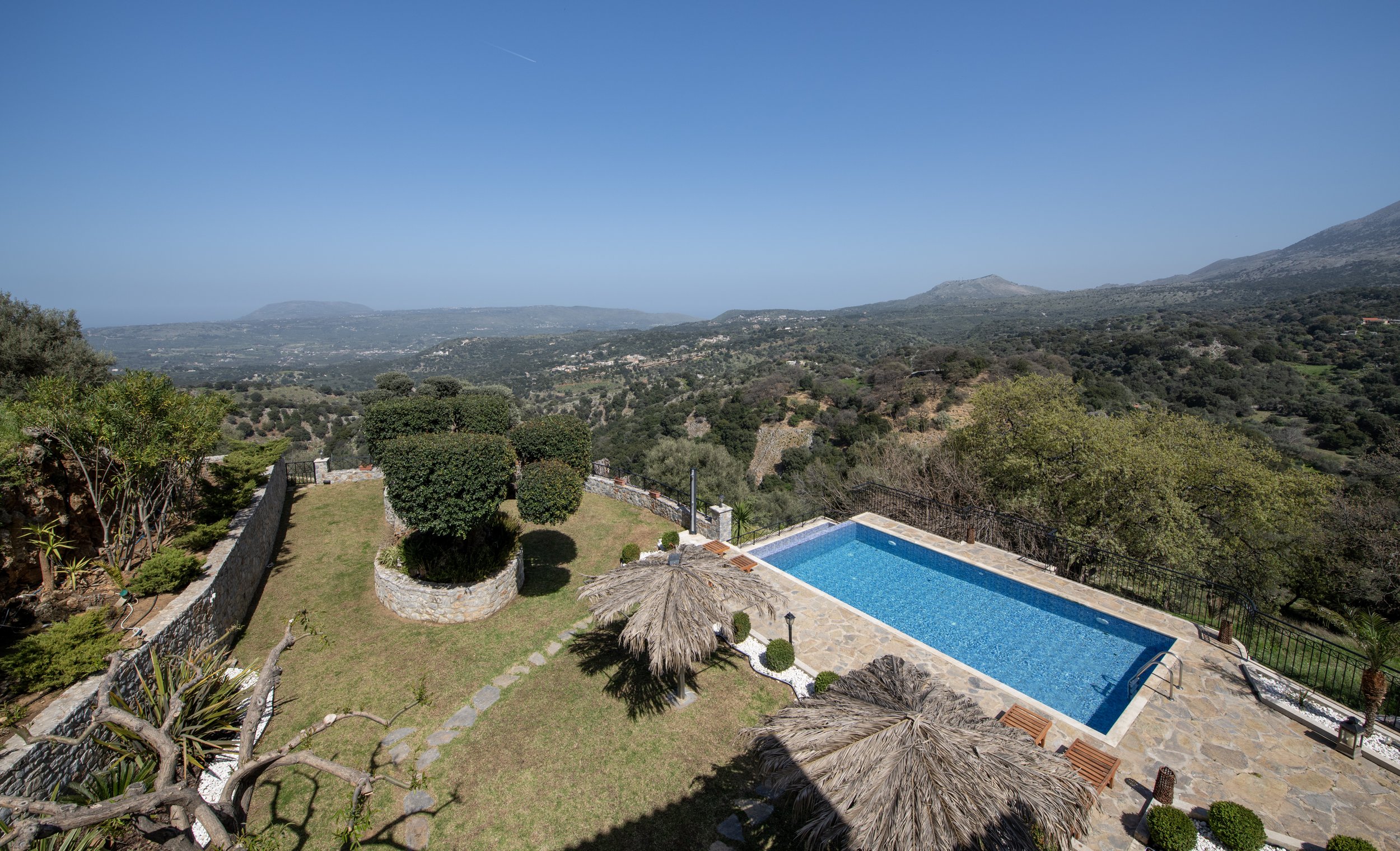 Aerial view of a serene landscape with a rectangular swimming pool, surrounded by a stone patio and lounge chairs with straw umbrellas. Lush green vegetation and mountainous terrain extend into the distance under a clear blue sky.