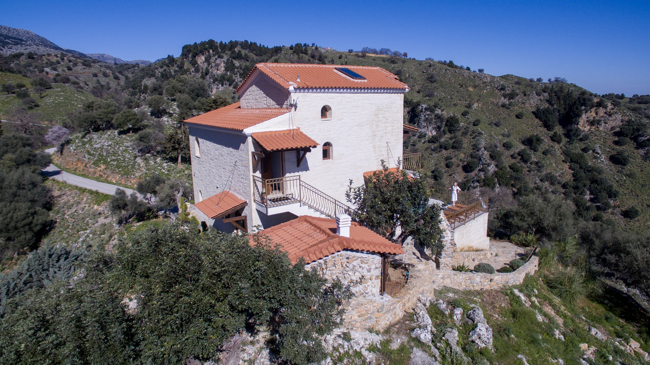 Stone house with red-tiled roof on a hillside surrounded by greenery and mountains.