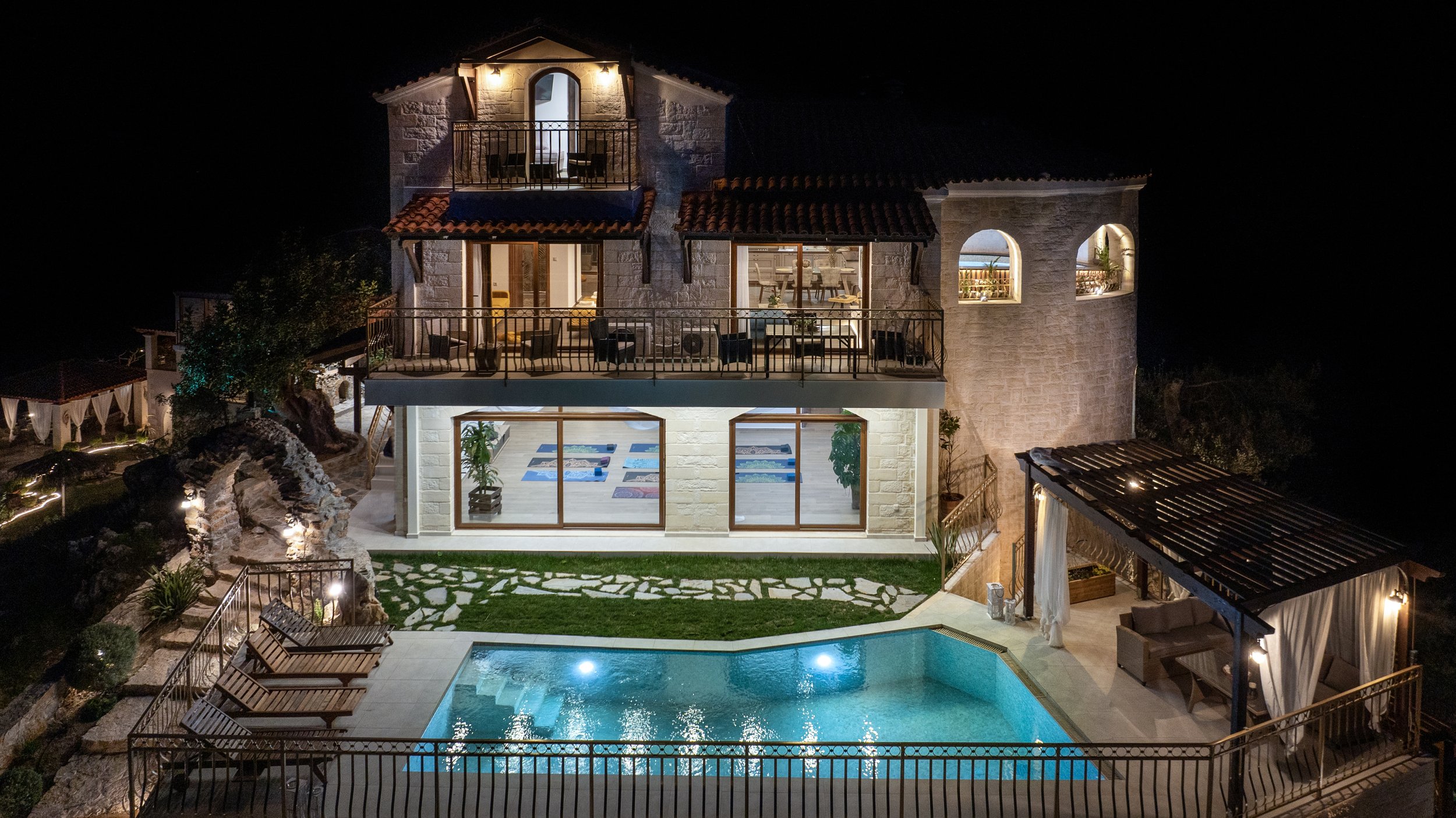 Luxurious multi-story villa at night with illuminated swimming pool, balconies, and outdoor seating.