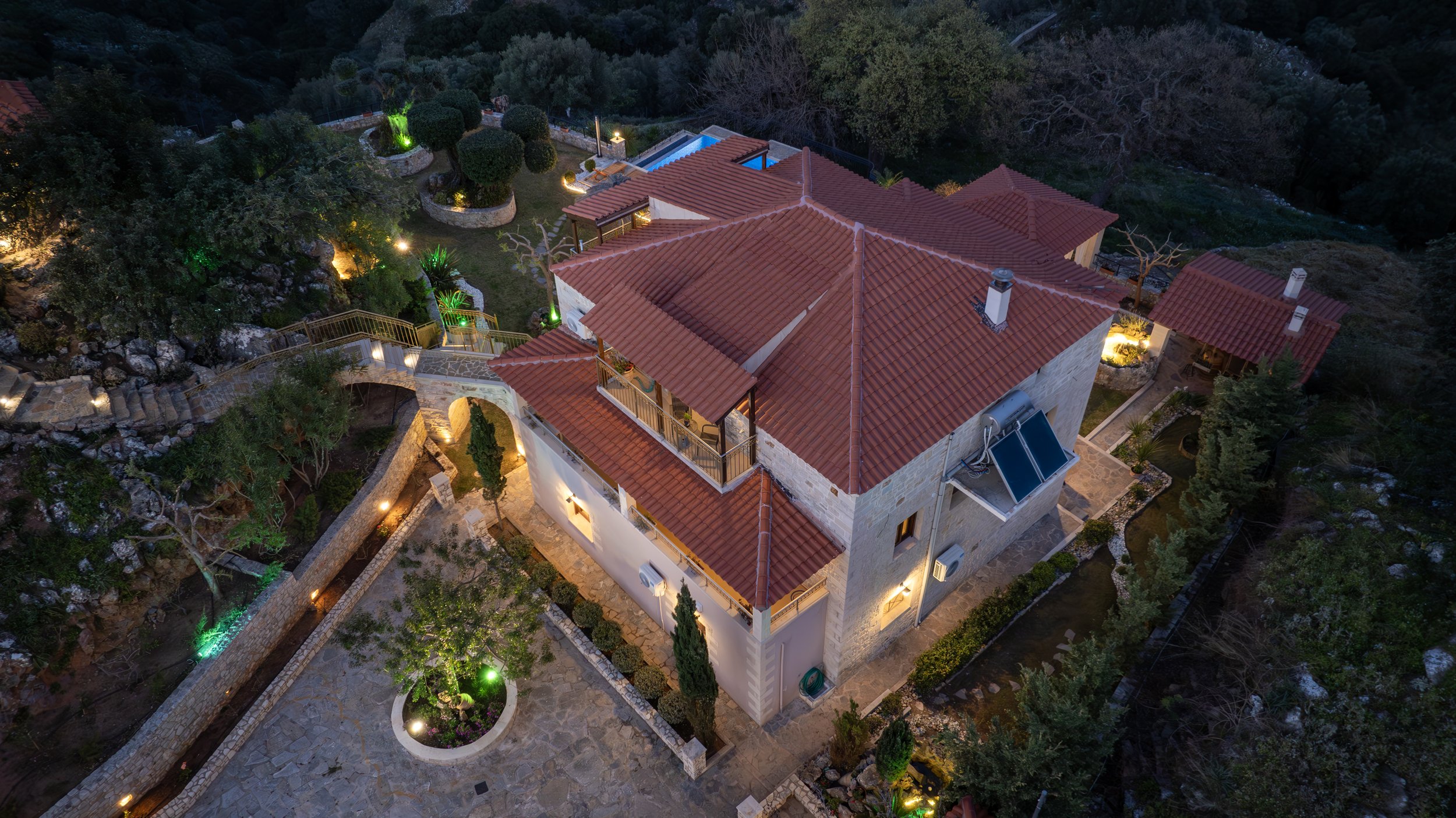 Aerial view of a stone house with red roof and surrounding garden, illuminated at night with pathways and decorative landscaping.