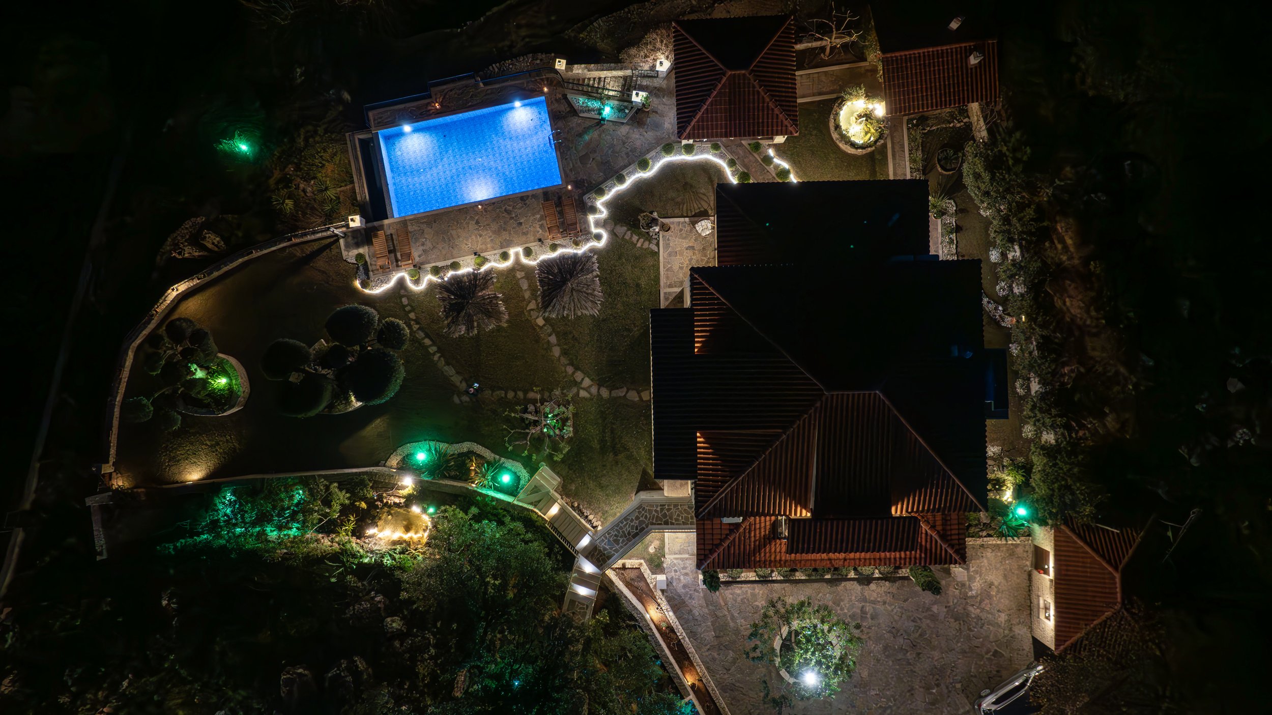 Aerial nighttime view of a house with an illuminated swimming pool, garden paths lined with lights, and surrounding landscaping.