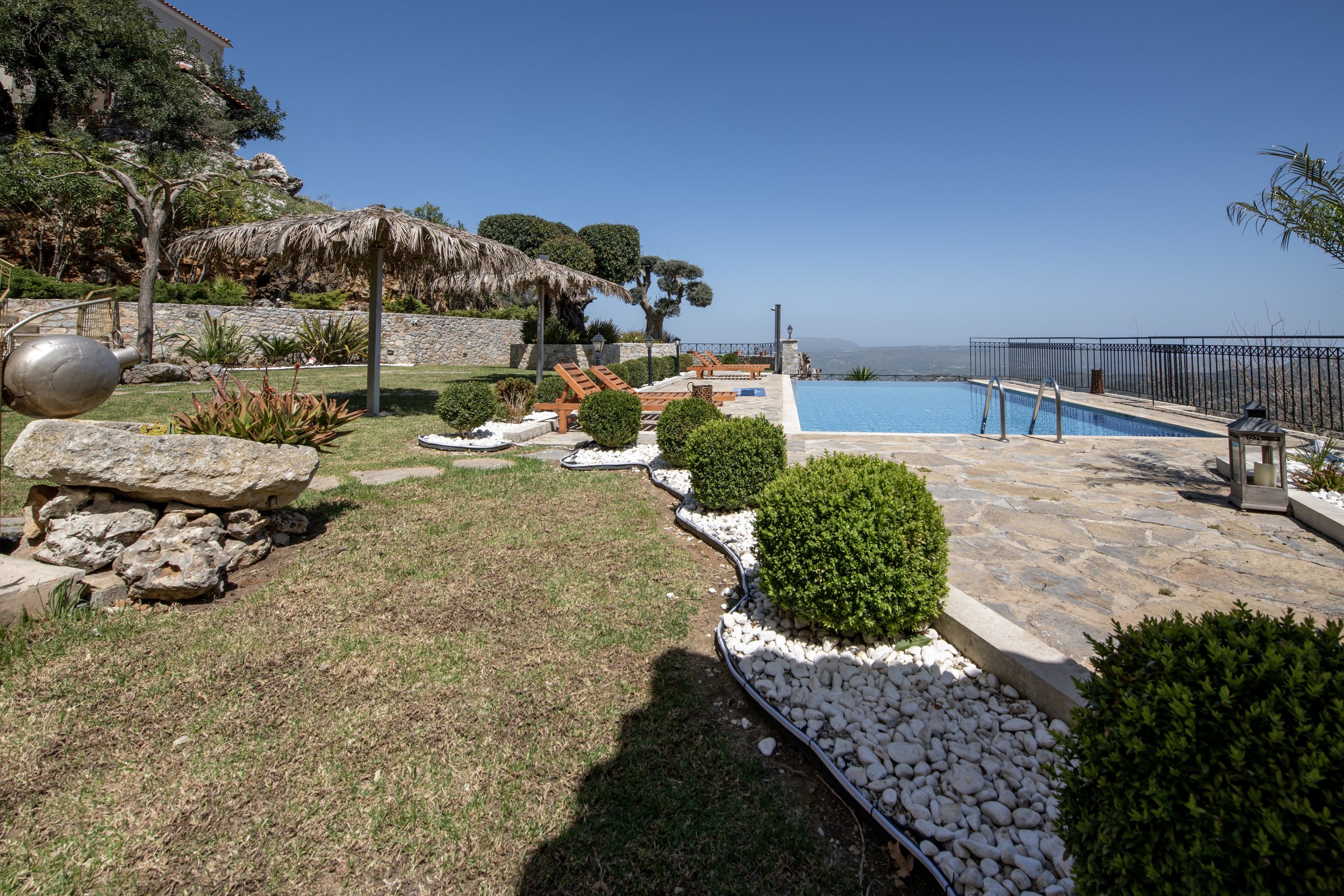 Outdoor pool area with a stone patio, sun loungers, a thatched umbrella, manicured bushes, and scenic view.