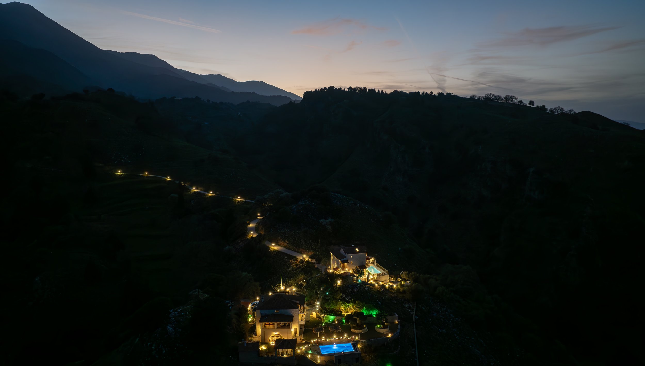 Aerial view of a hillside landscape at dusk with illuminated buildings and a winding road.