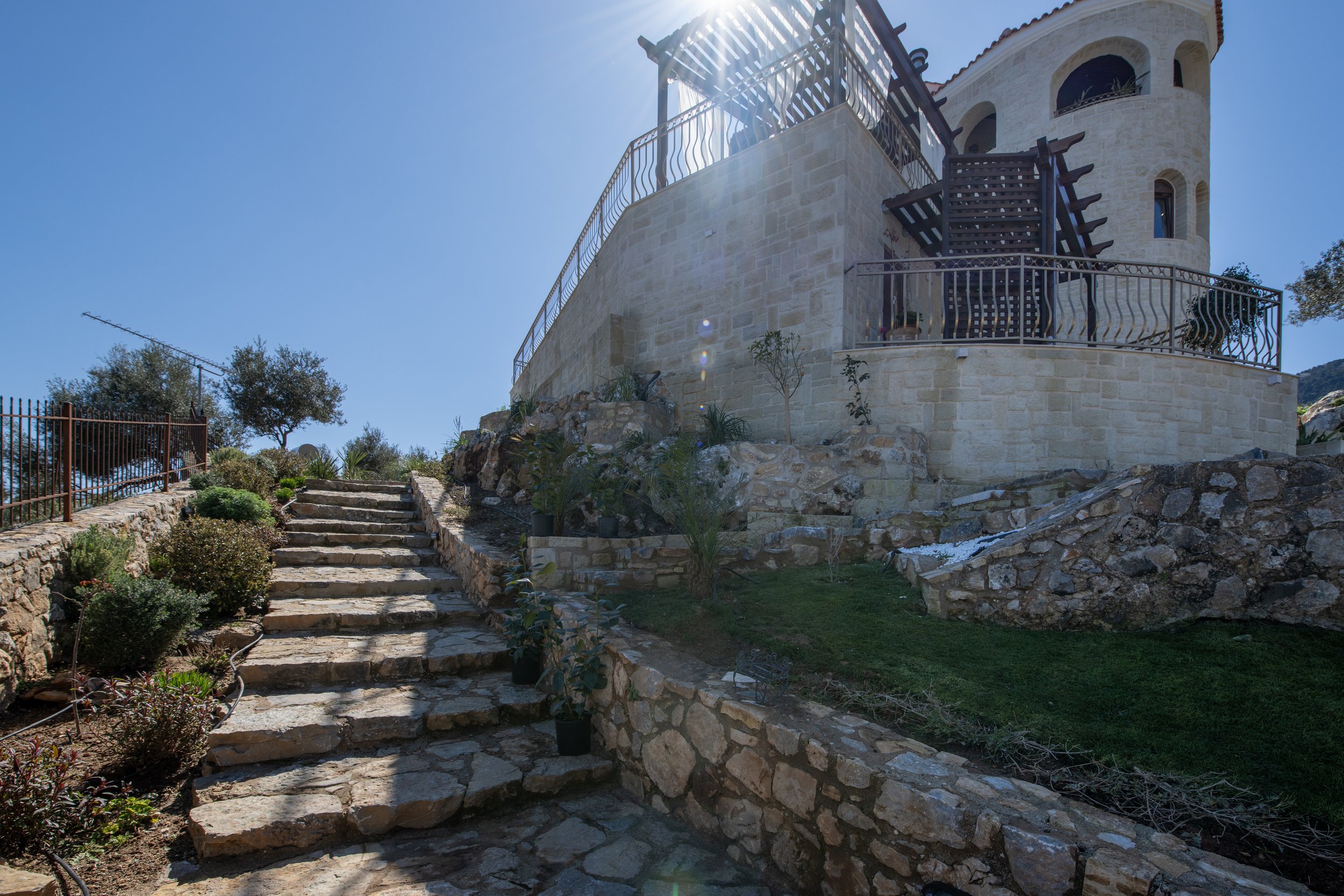 Stone staircase leading to a villa with a pergola, surrounded by landscaped gardens and plants, under a clear blue sky.
