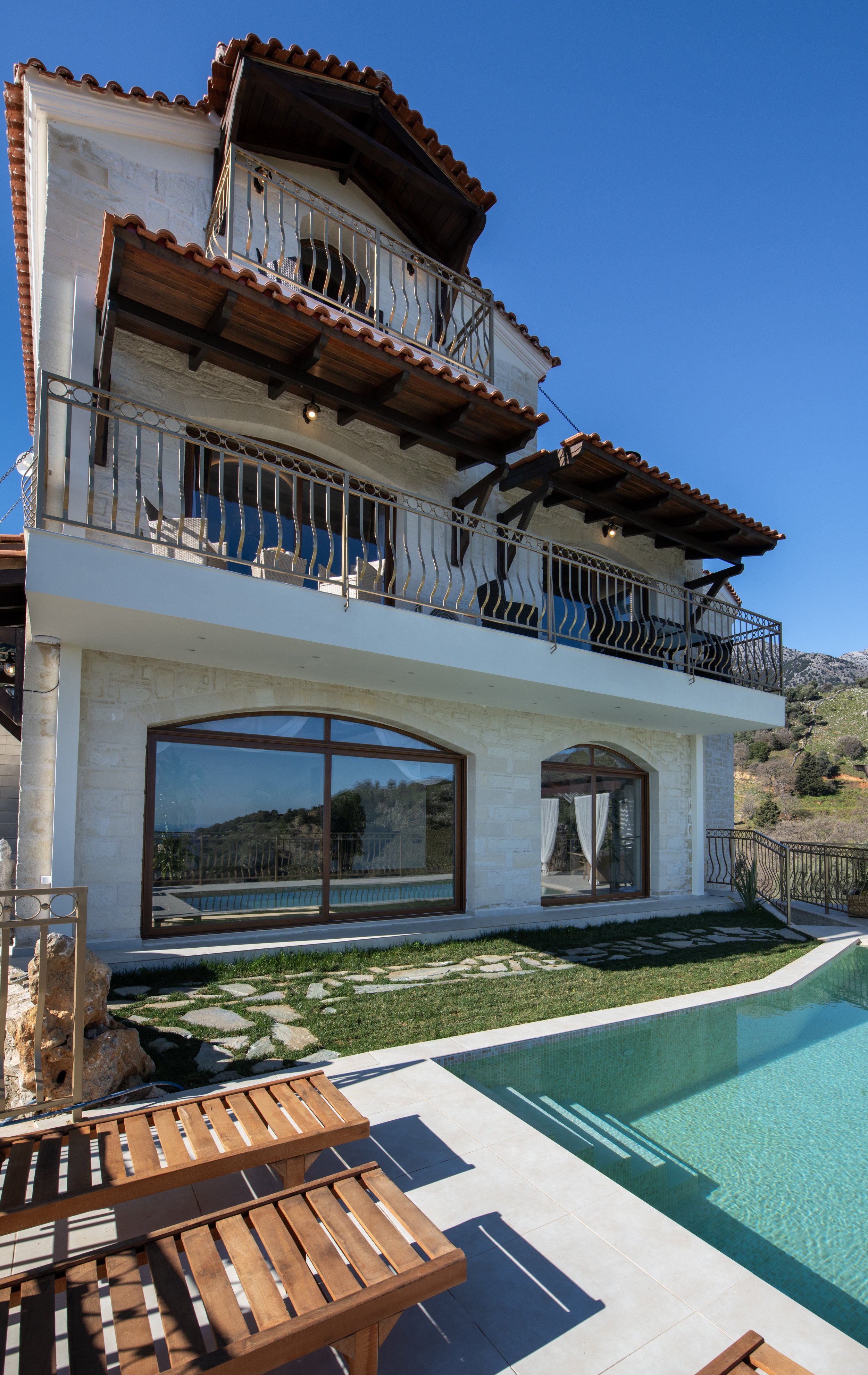 Exterior view of a Mediterranean-style house with balconies, stone walls, and a swimming pool in the foreground. The sky is clear and blue.