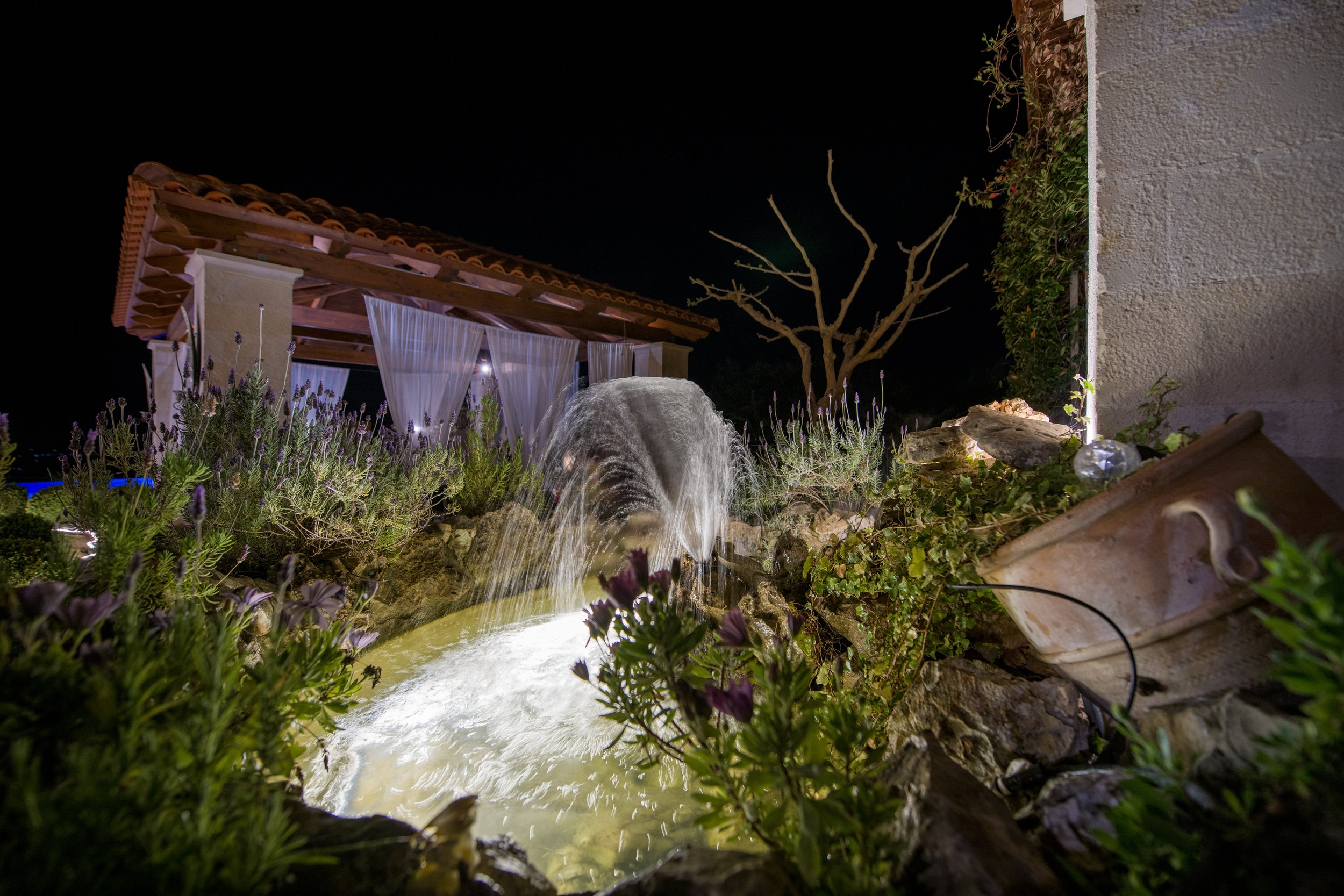 Outdoor night scene with fountain, stone archway, and greenery