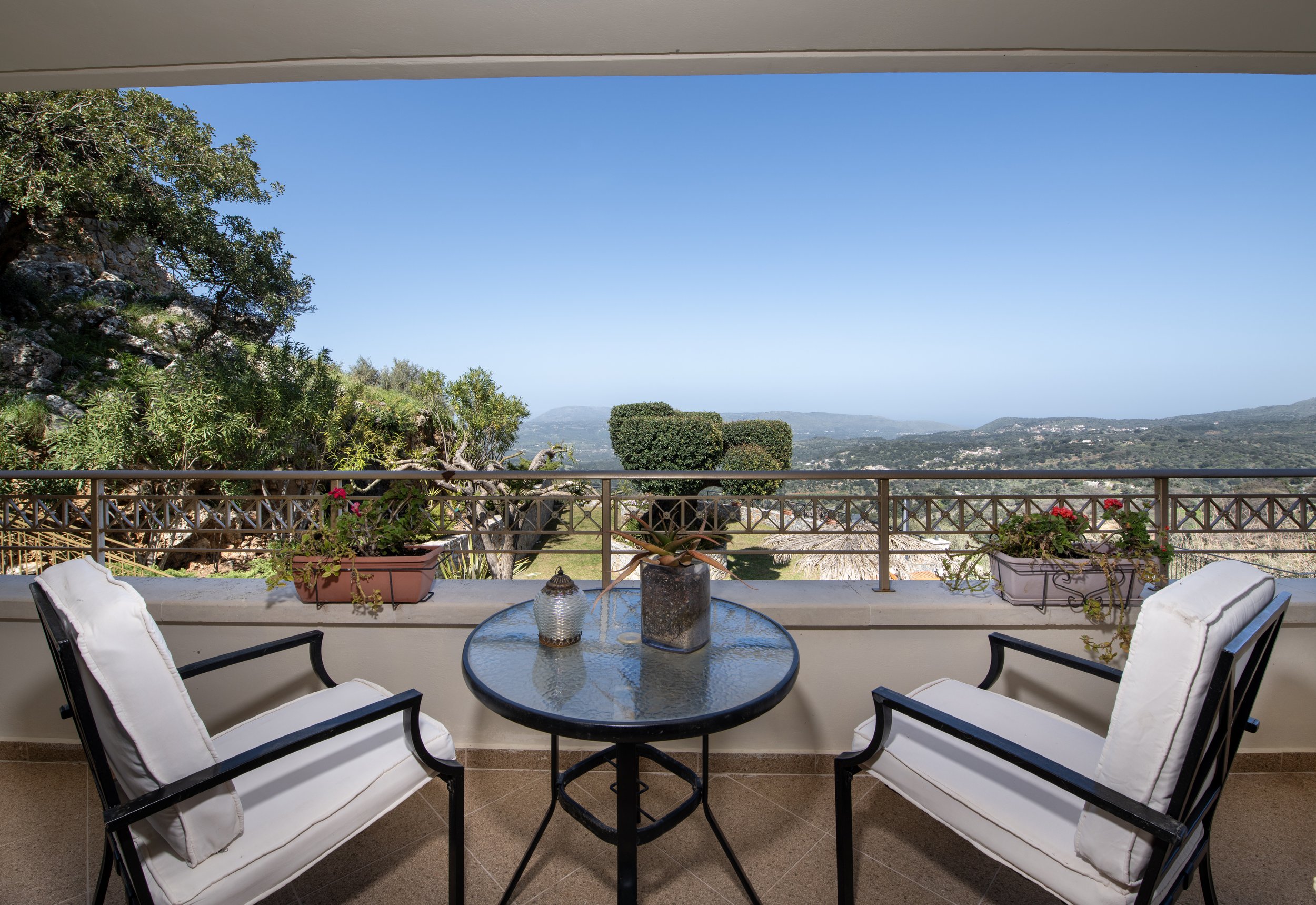 Terrace with two chairs and a glass table, overlooking a scenic valley with hills and greenery, framed by trees and potted plants.