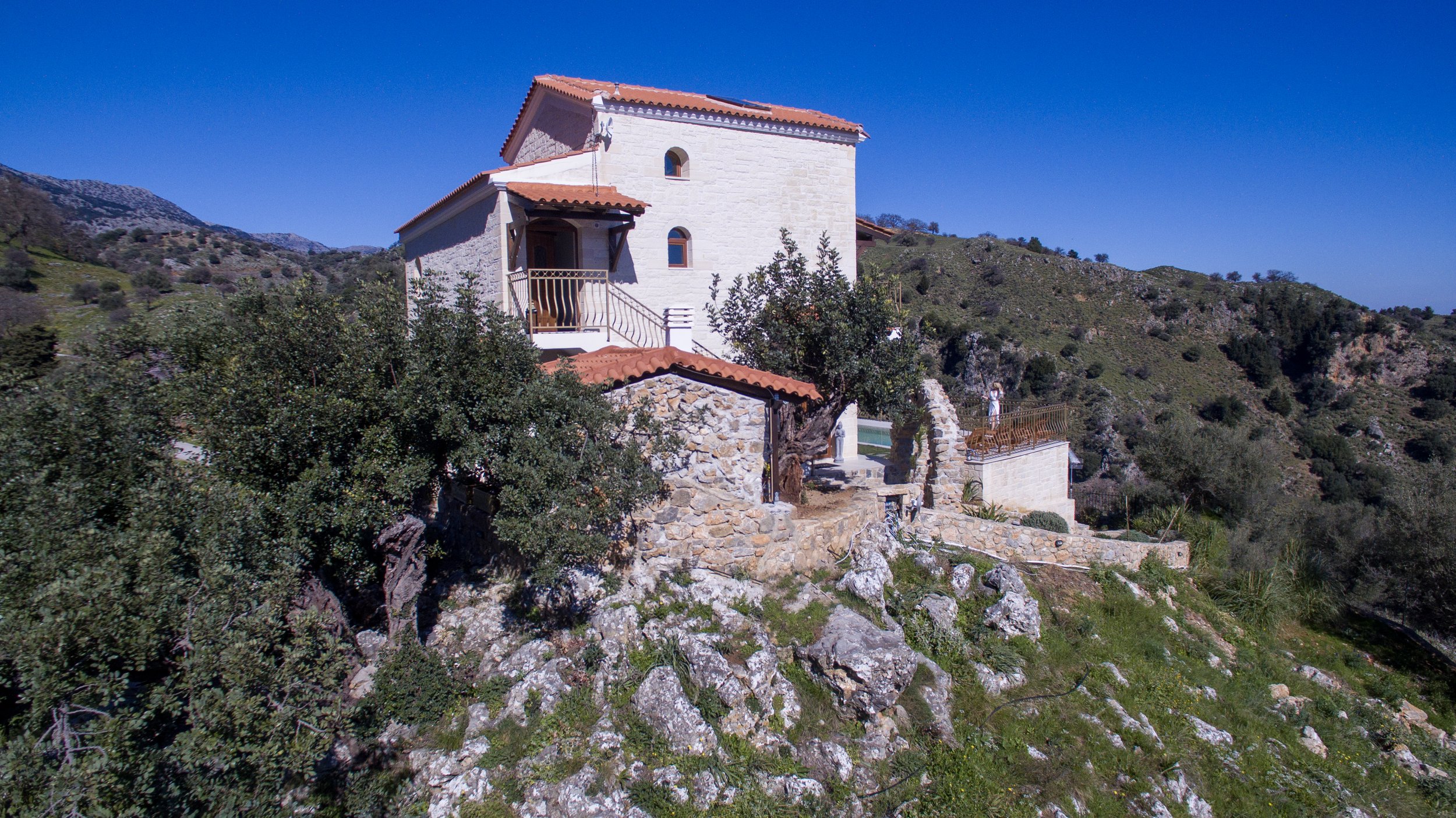 A stone house with a red-tiled roof situated on a hillside surrounded by greenery under a clear blue sky.
