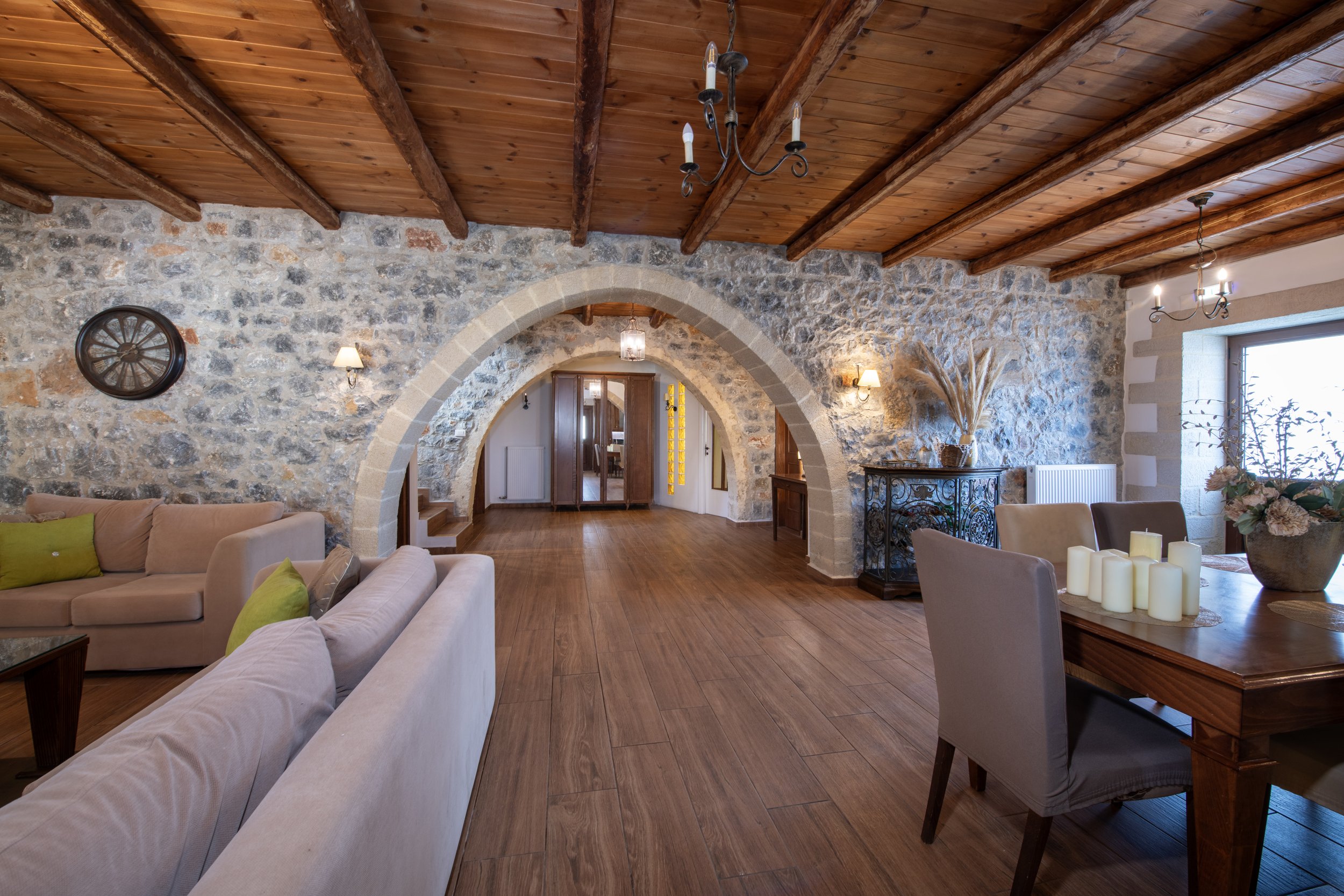 Rustic living room with stone walls, wooden ceiling beams, arched entrances, beige sofas with green pillows, dining area with candles, and decorative plants.