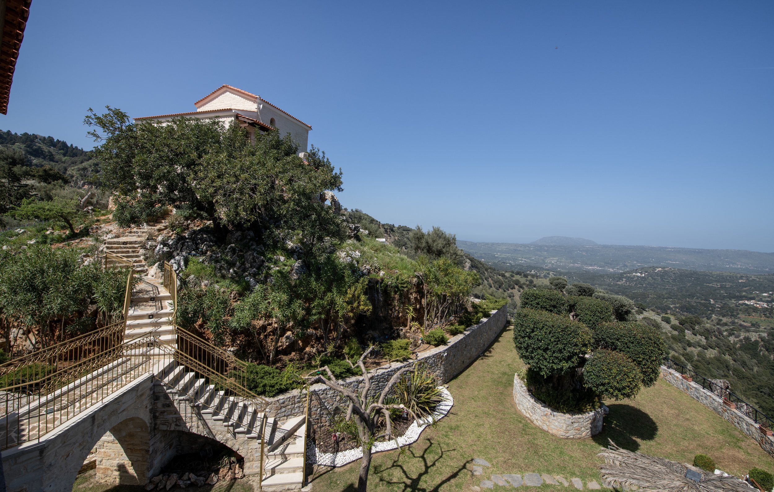 Hilltop house with stone staircase, surrounded by lush greenery, overlooking a scenic valley with distant mountains under a clear blue sky.