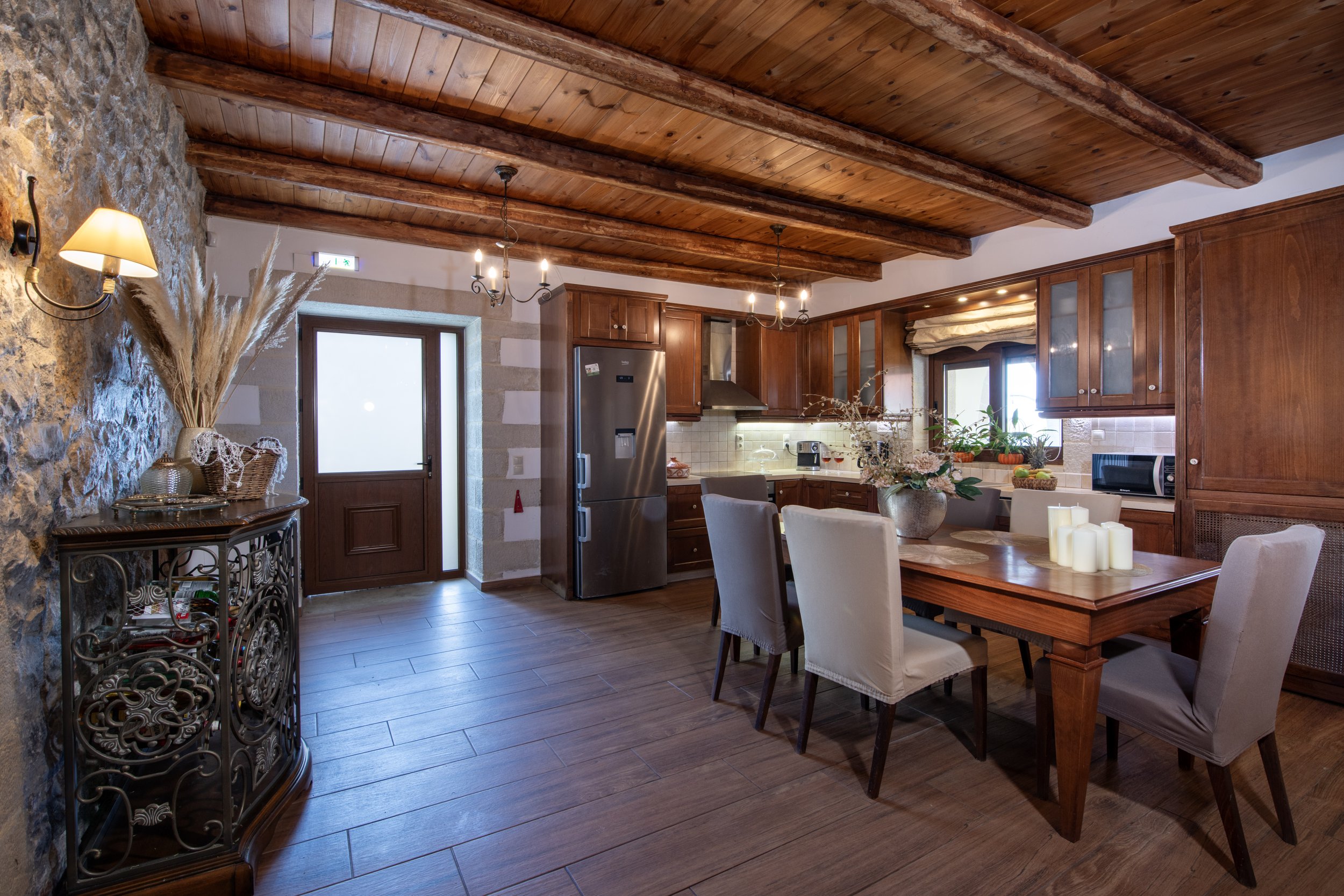 Rustic kitchen interior with wooden beams, stone wall, wooden cabinets, dining table with chairs, stainless steel refrigerator, and decorative elements like plants, candles, and a vase with dried flowers.