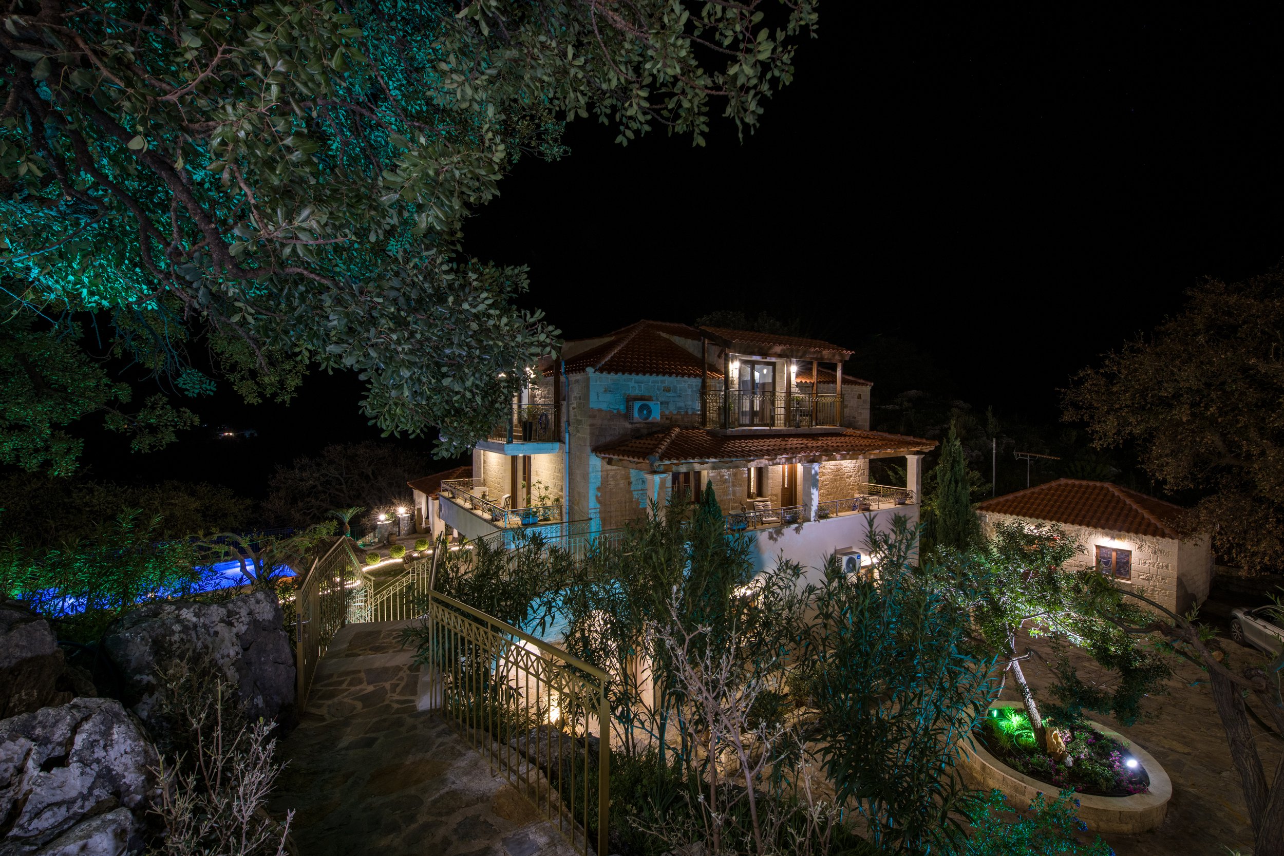 Mediterranean-style villa with stone walls, illuminated at night, surrounded by trees and plants. A stone path and railings lead to the main entrance.