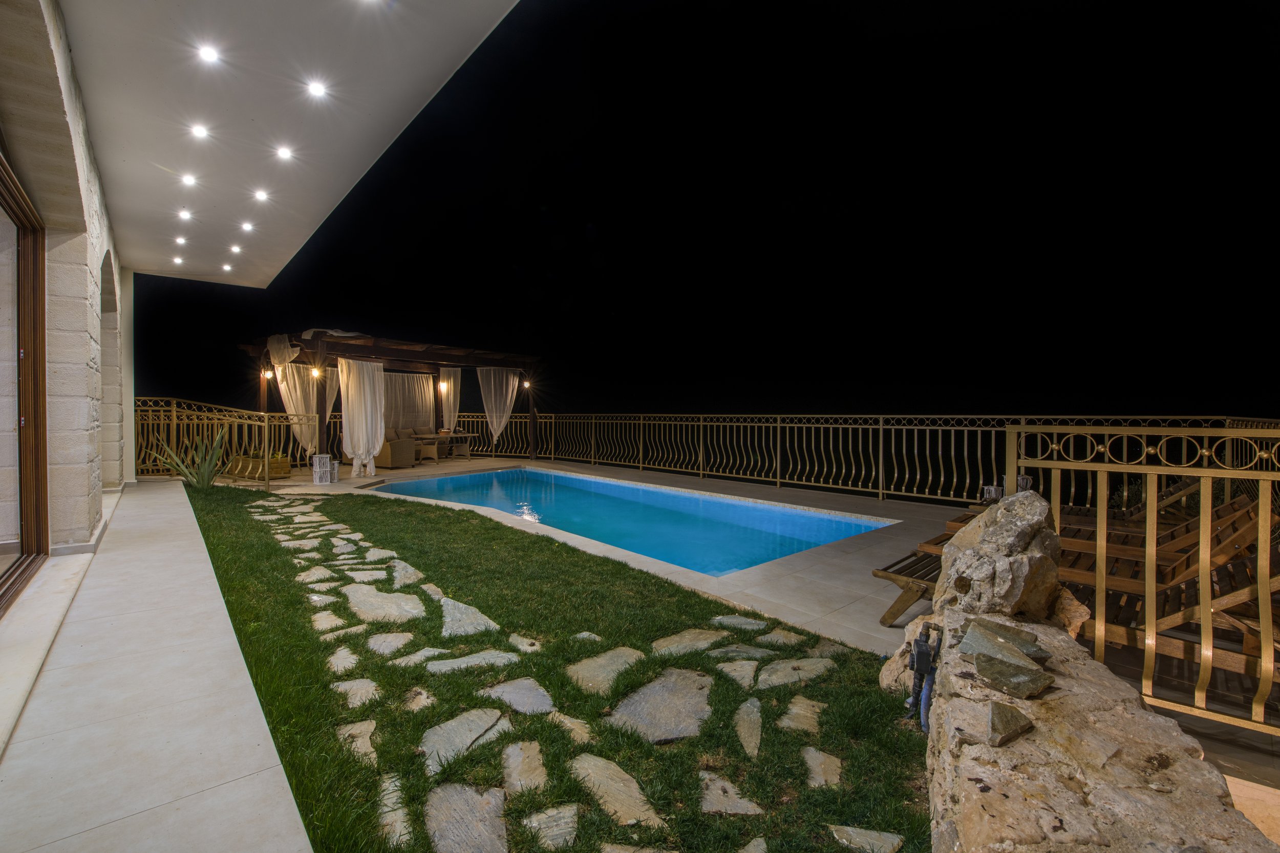 Outdoor patio at night with illuminated swimming pool, stone pathway, and seating area under a pergola with curtains.