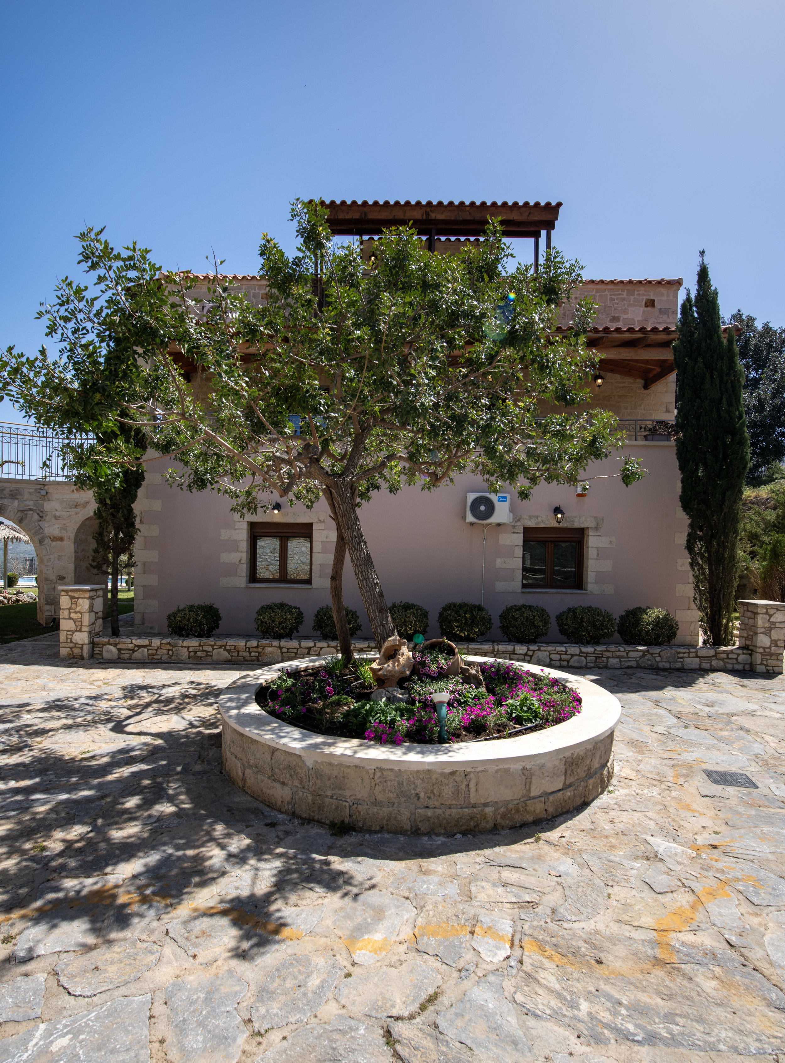 A Mediterranean-style stone house with a tile roof, featuring a circular stone planter with a tree and purple flowers in the courtyard on a sunny day.