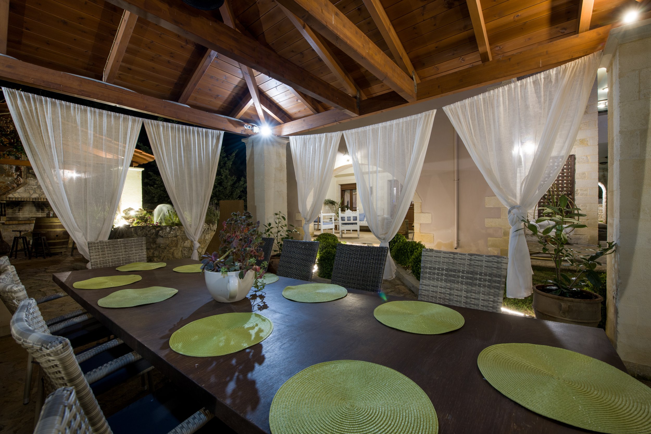 Outdoor dining area with a large wooden table, wicker chairs, and green placemats. The space features sheer white curtains, wooden ceiling beams, and ambient lighting.