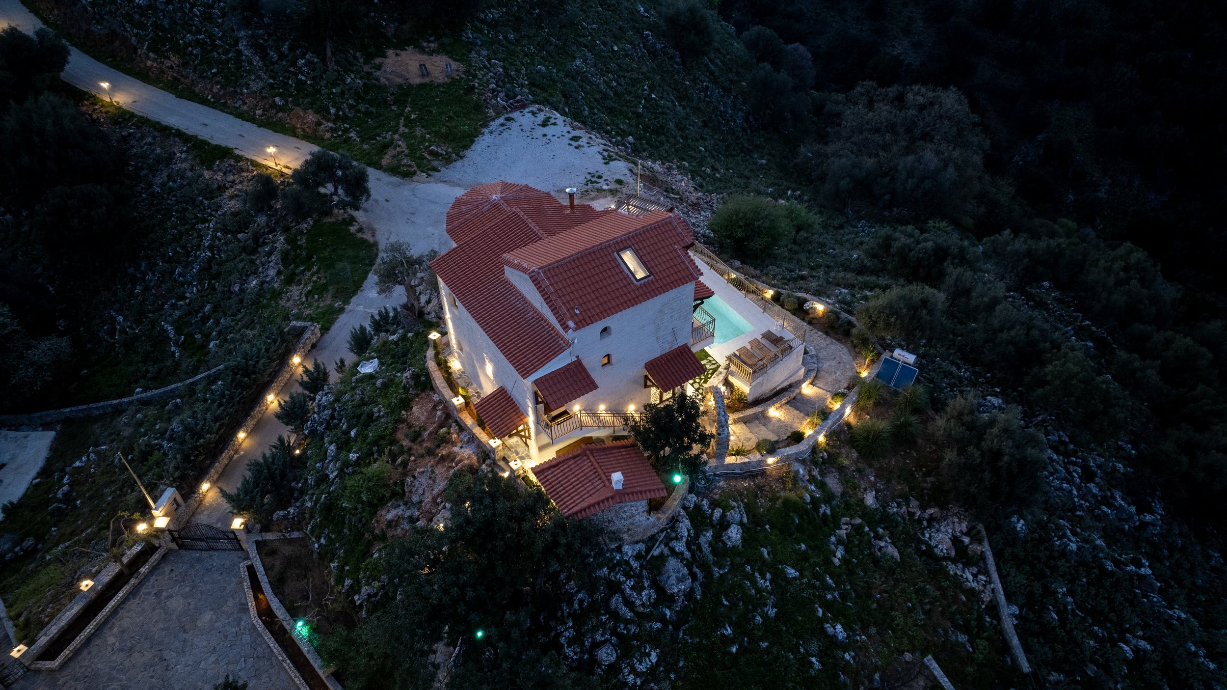 Aerial view of a house with terracotta roof and outdoor pool, illuminated at night, surrounded by trees and landscape.