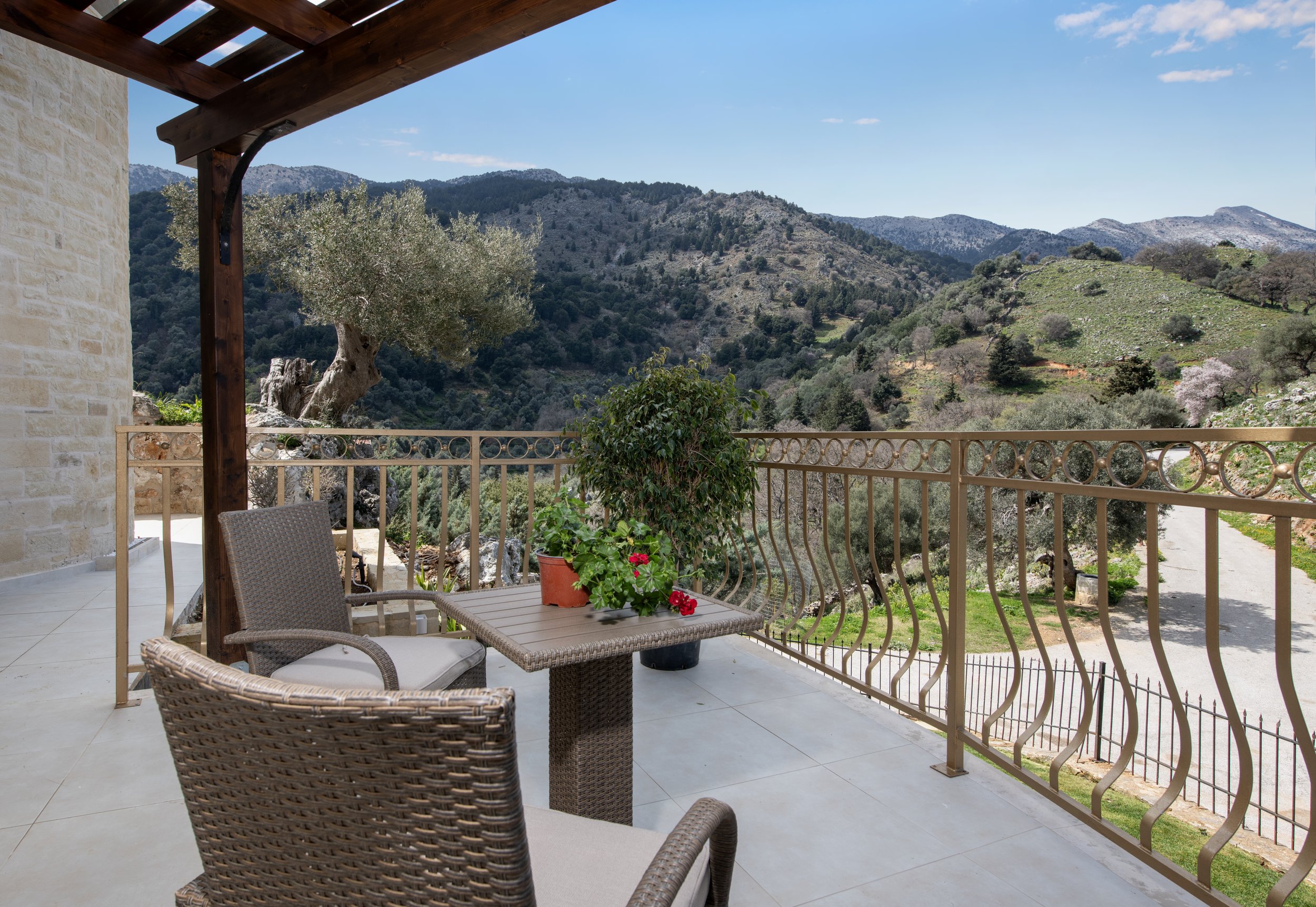 Mountain view from a balcony with wicker chairs and table, potted plants, and iron railing.