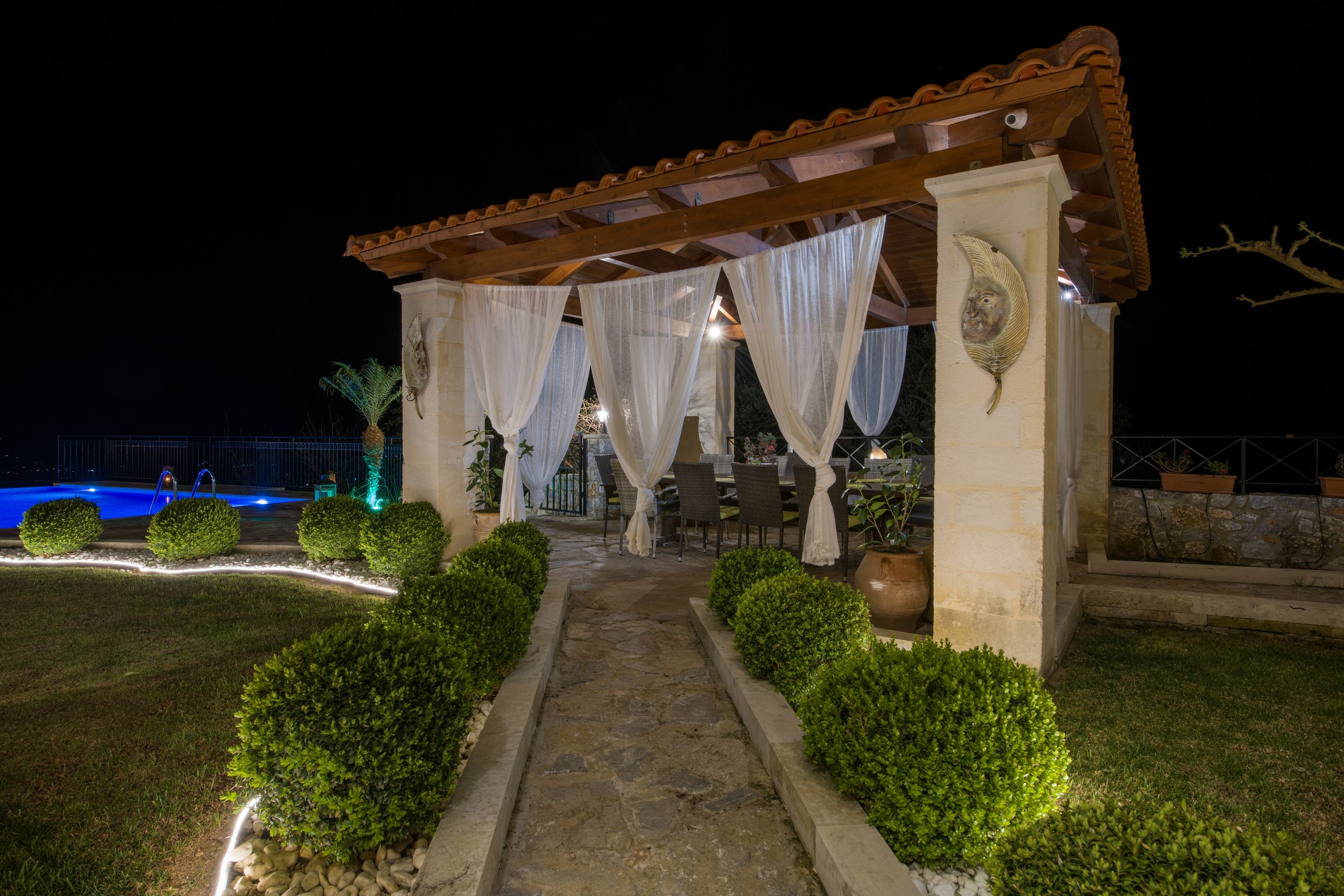 Outdoor gazebo with curtains and stone path at night, surrounded by plants and illuminated by lights.