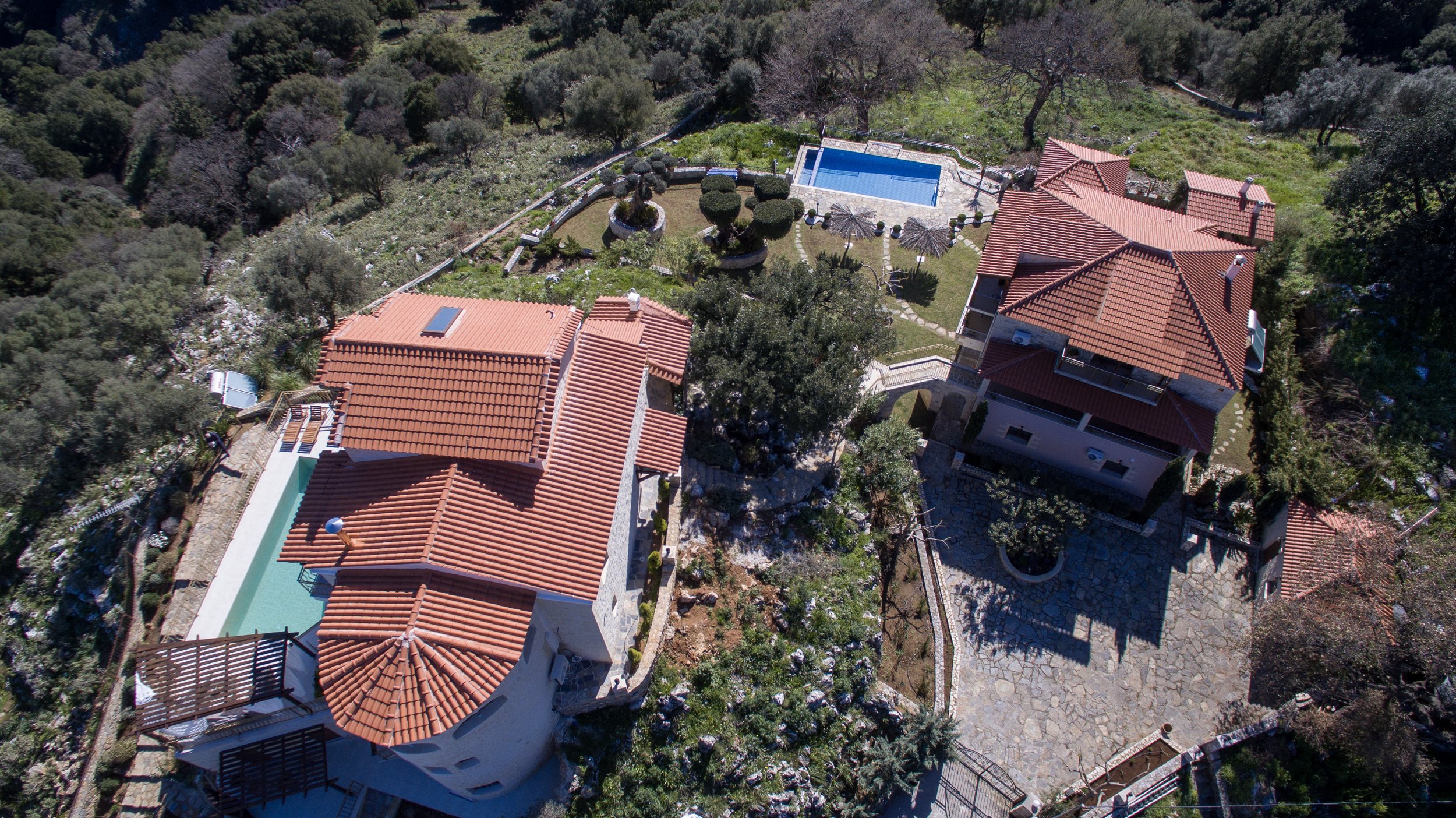 Aerial view of two houses with red-tiled roofs, a swimming pool, and landscaped gardens surrounded by trees.
