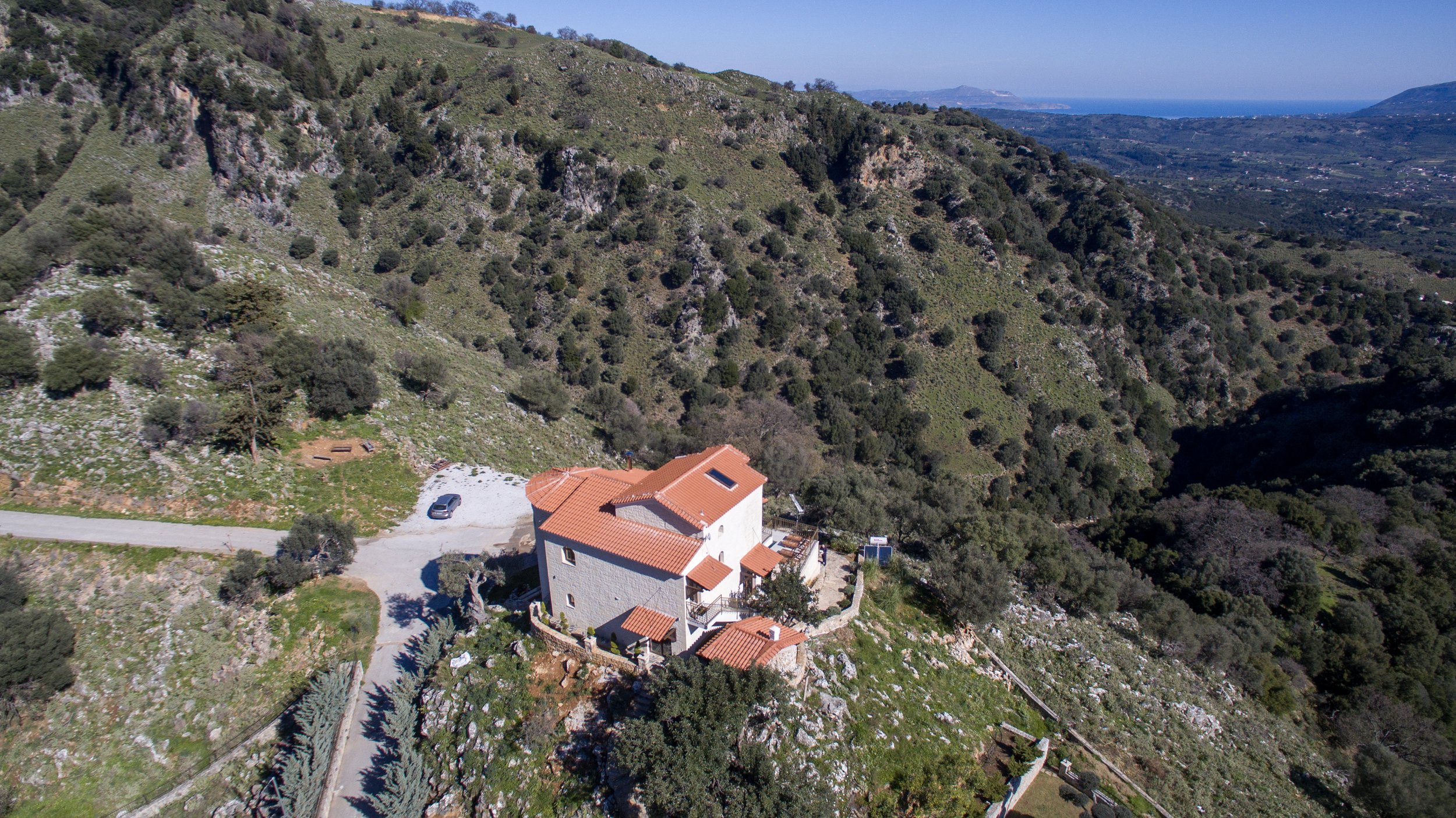 Aerial view of a house with a red-tiled roof situated on a hillside surrounded by green vegetation and rocky terrain. A road and a parked car are visible, with expansive landscape views in the background, including distant hills and a glimpse of the