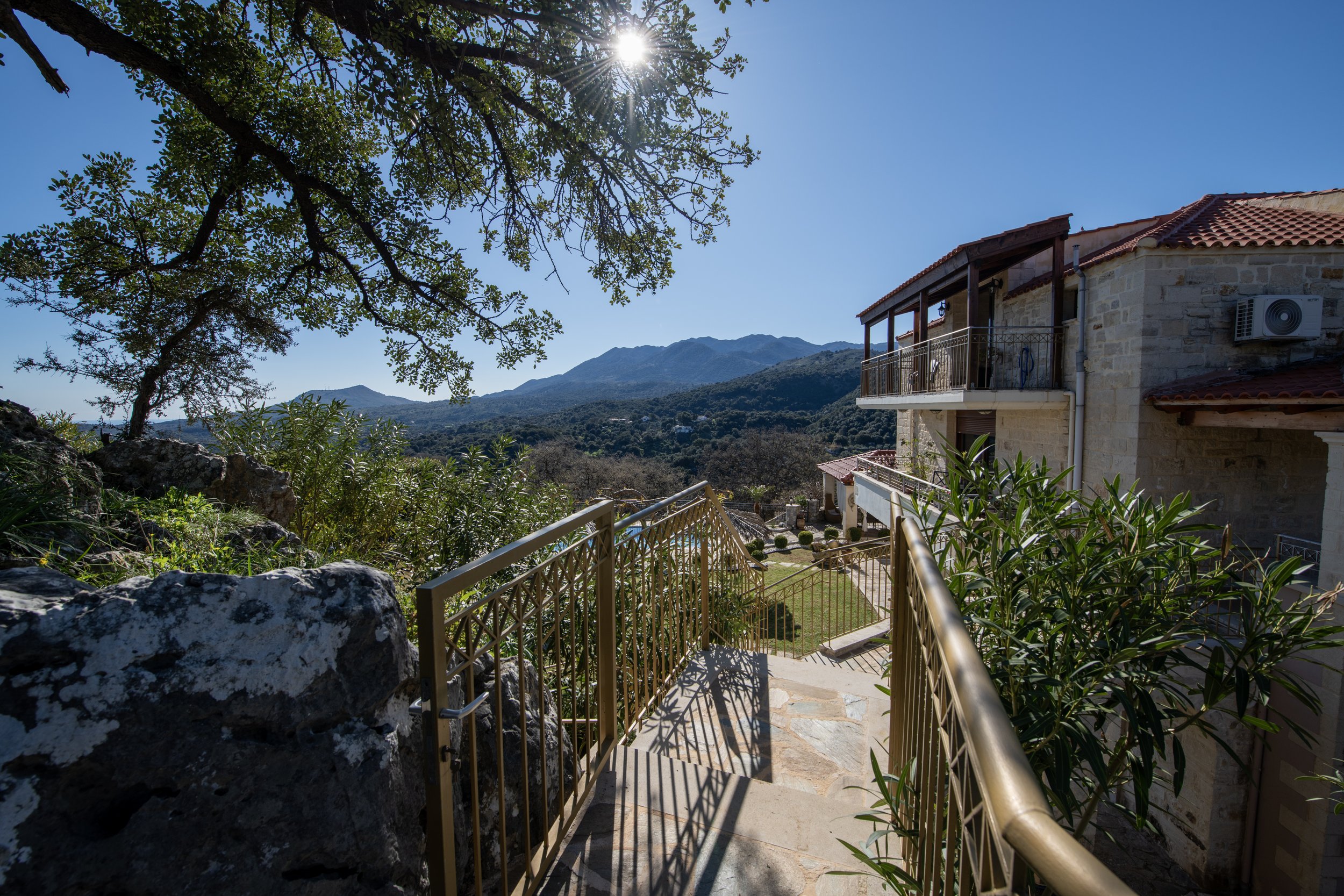 Sunny landscape with a stone house, metal railings, and a tree in the foreground; mountains in the background.