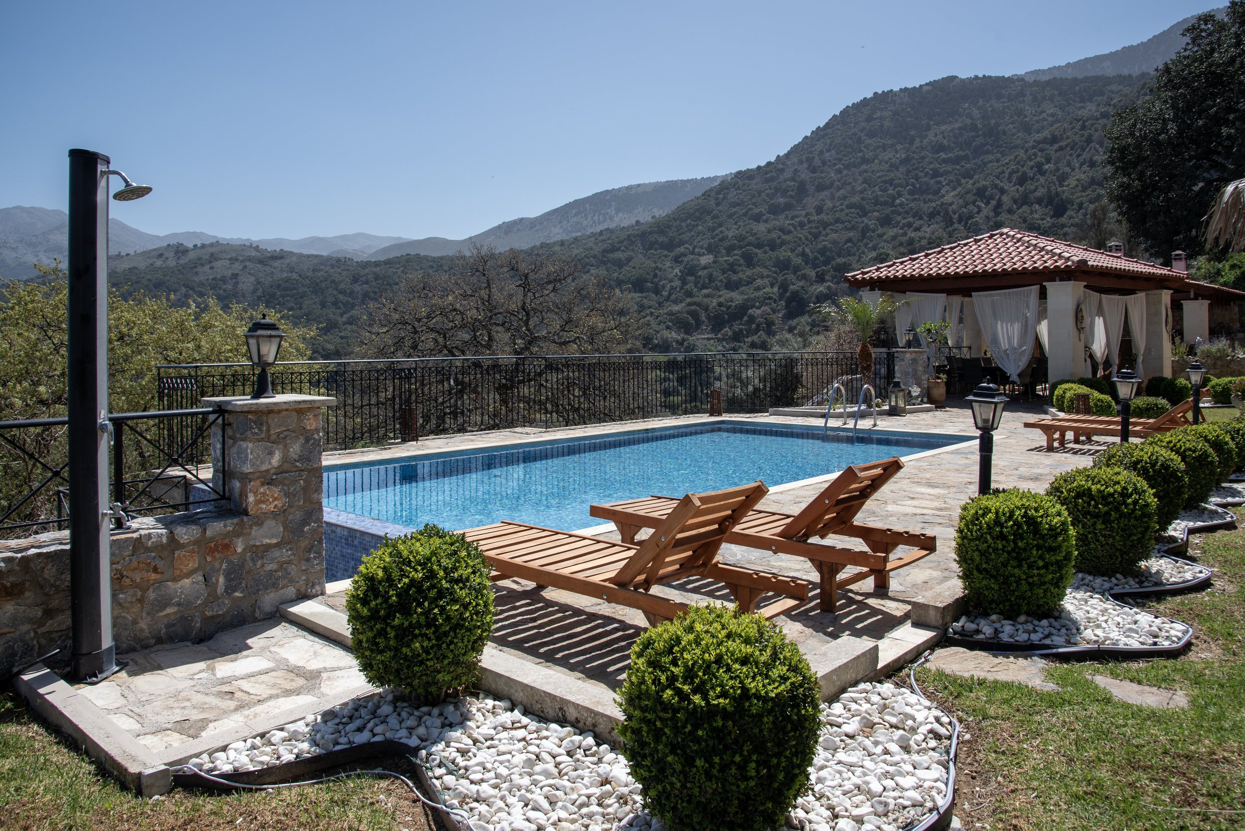 Outdoor pool area with wooden lounge chairs, stone pathway, decorative shrubbery, a gazebo with white curtains, and a scenic mountain view in the background.