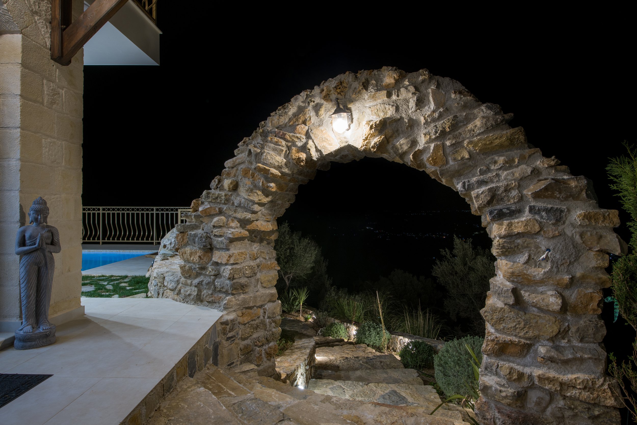 Stone arch and statue by a pool at night, with trees and city lights in the background.