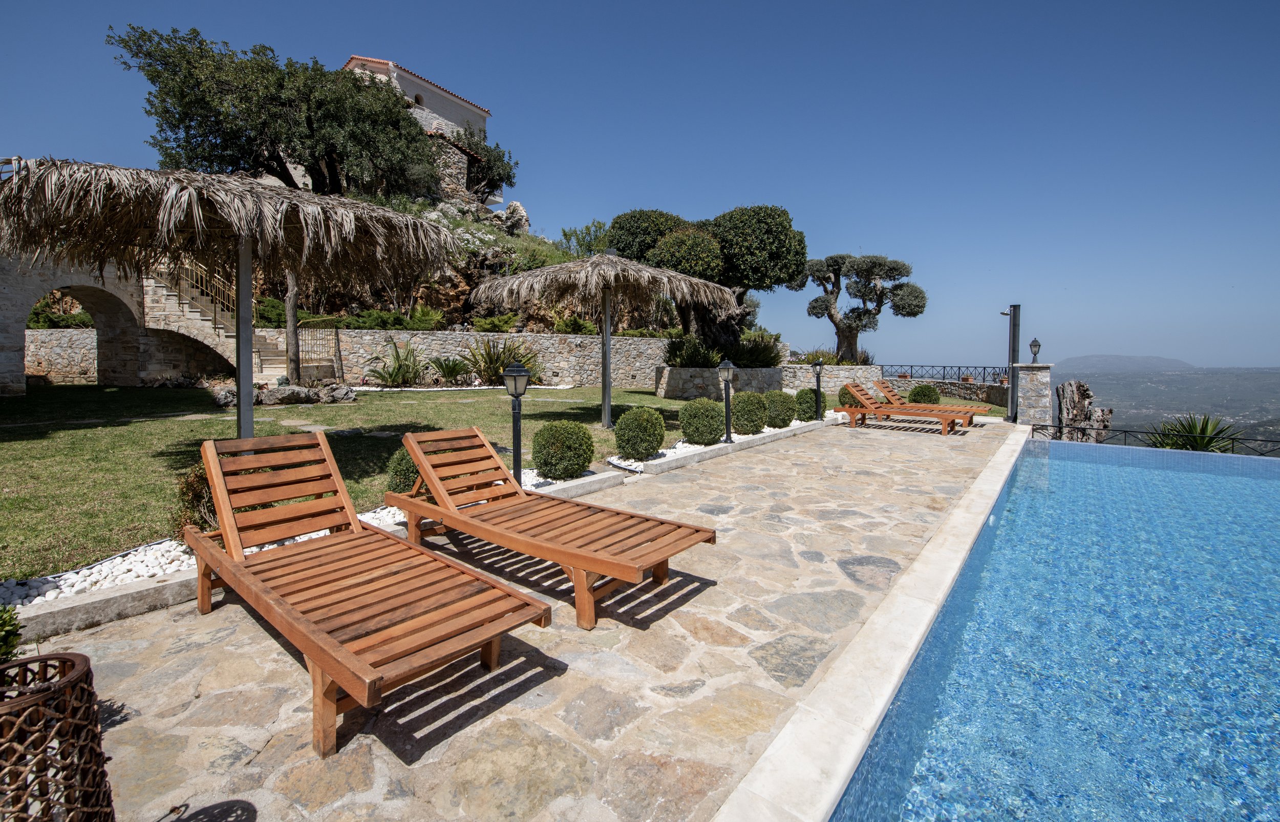 Outdoor pool area with wooden lounge chairs, stone patio, grass, and a rustic stone building with trees in the background.