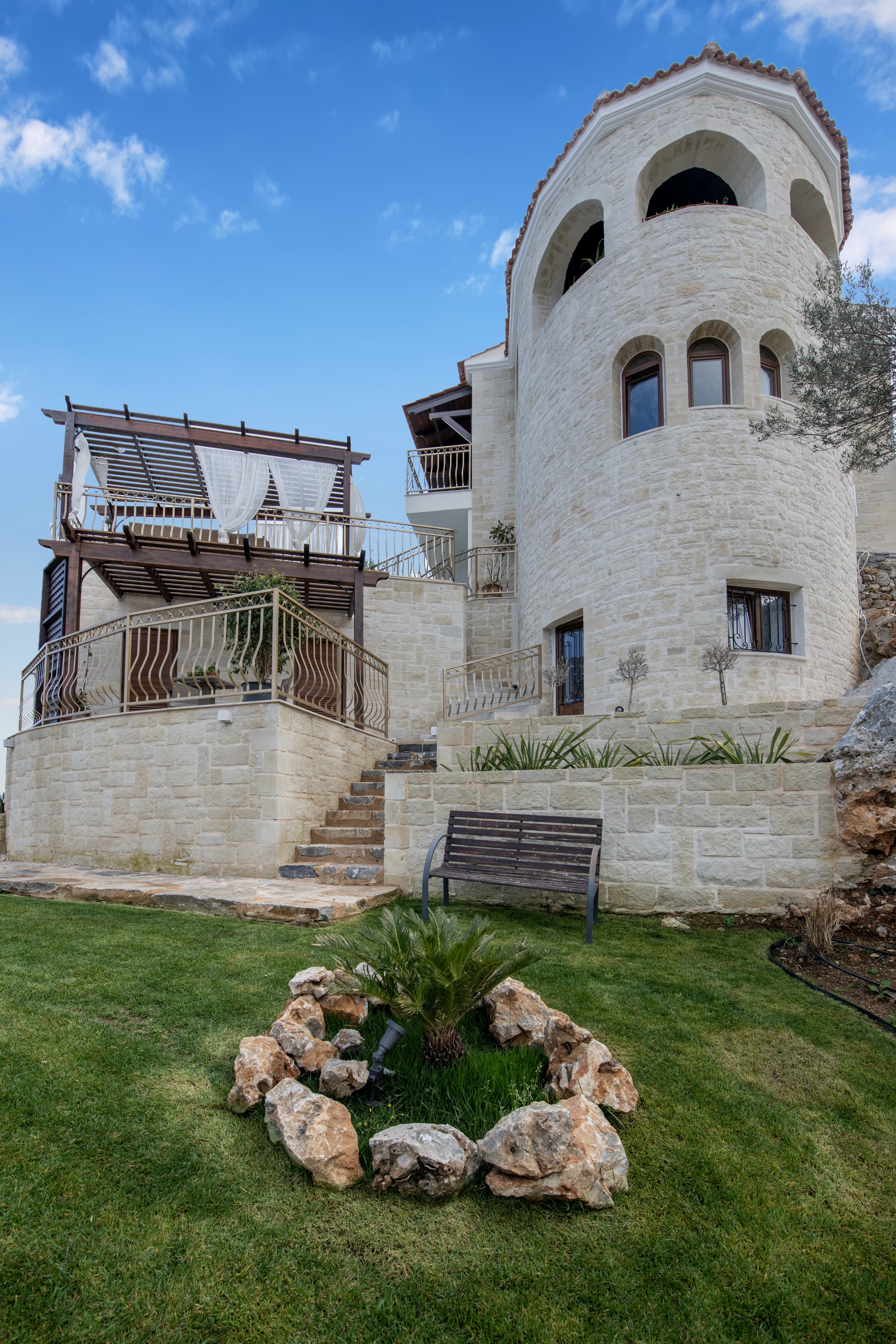 Mediterranean-style stone villa with arched windows, staircase, pergola, and landscaped garden featuring a bench and rock-bordered plant area.