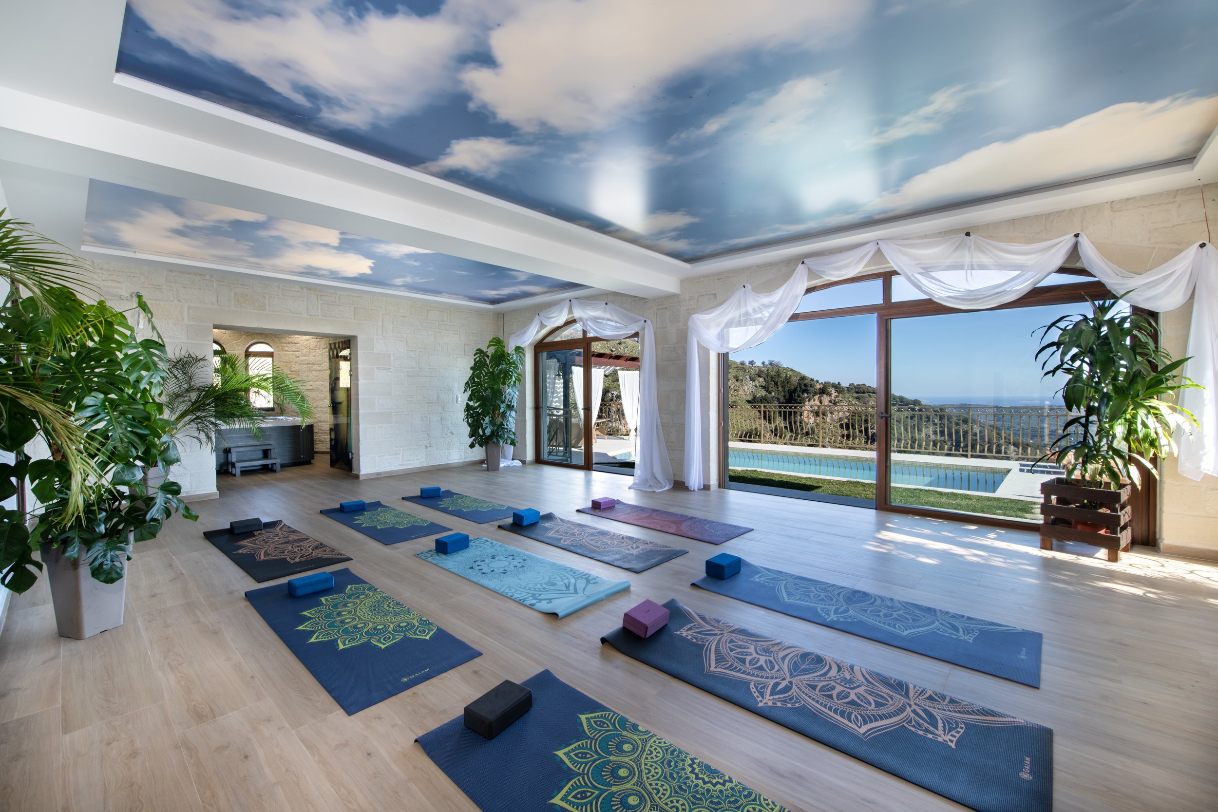 Yoga studio interior with decorative ceiling, multiple yoga mats, blocks, and large windows overlooking a pool and landscape.