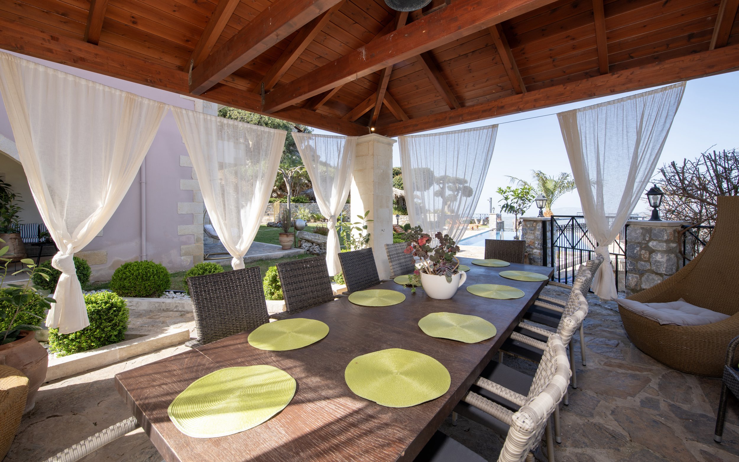 Outdoor patio area with a wooden table and wicker chairs under a wooden pergola, bordered by sheer white curtains. The table is set with green placemats and a white planter with a plant. Background includes a garden with small shrubs and a pool visib