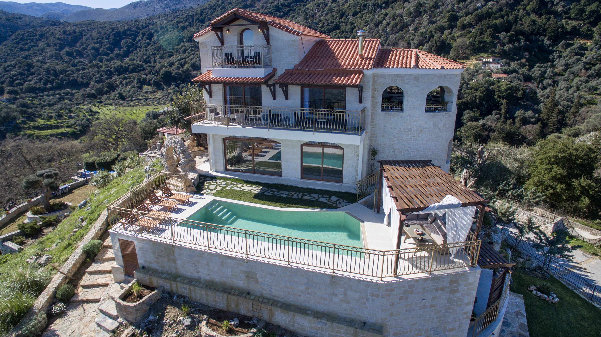 Luxury stone villa with a red-tiled roof, surrounded by lush green hills. Features include a swimming pool with sun loungers, multiple balconies, and a pergola-covered seating area, providing expansive views of the scenic landscape.