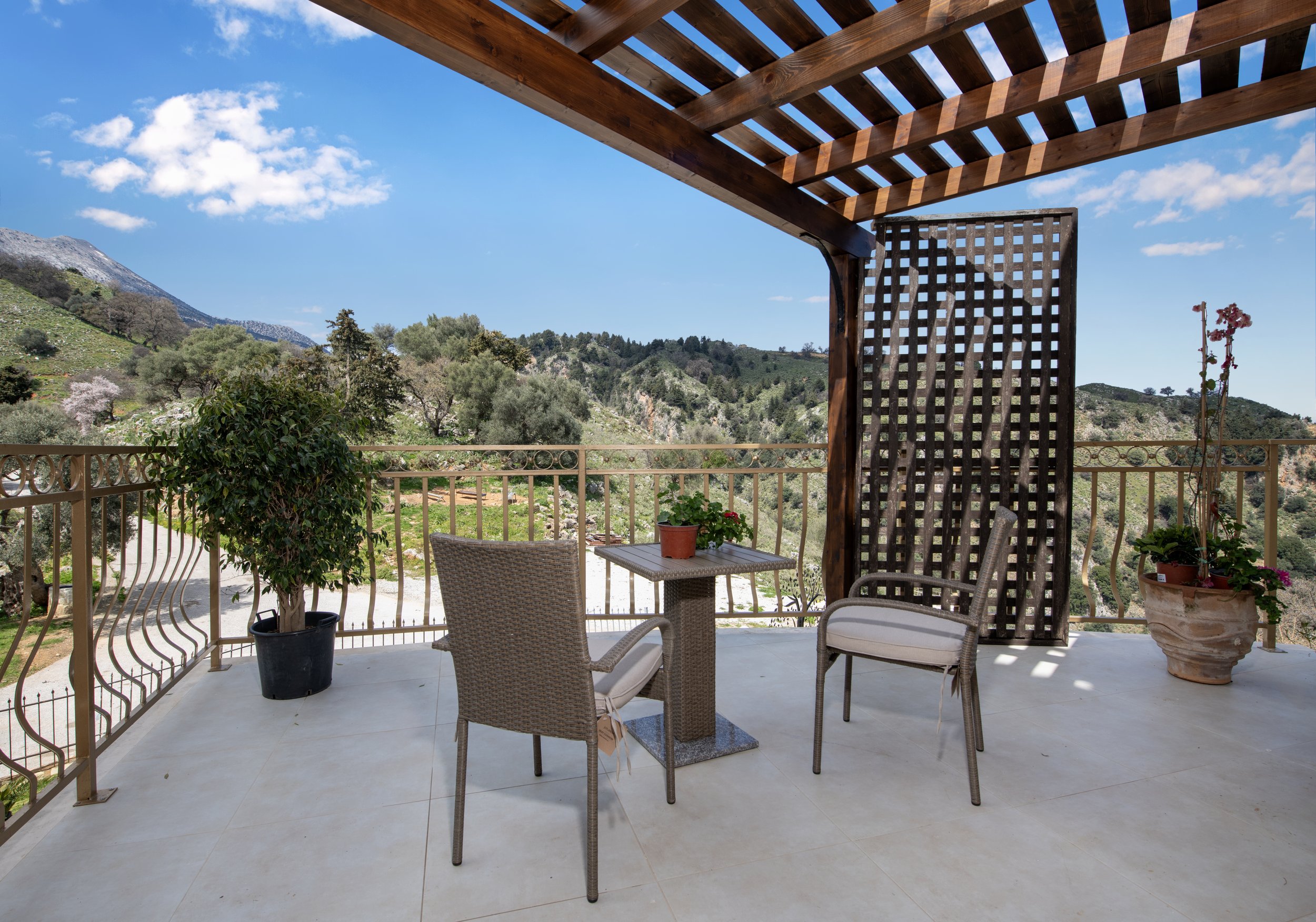Outdoor patio with chairs, table, potted plants, and wooden pergola overlooking mountains and greenery.
