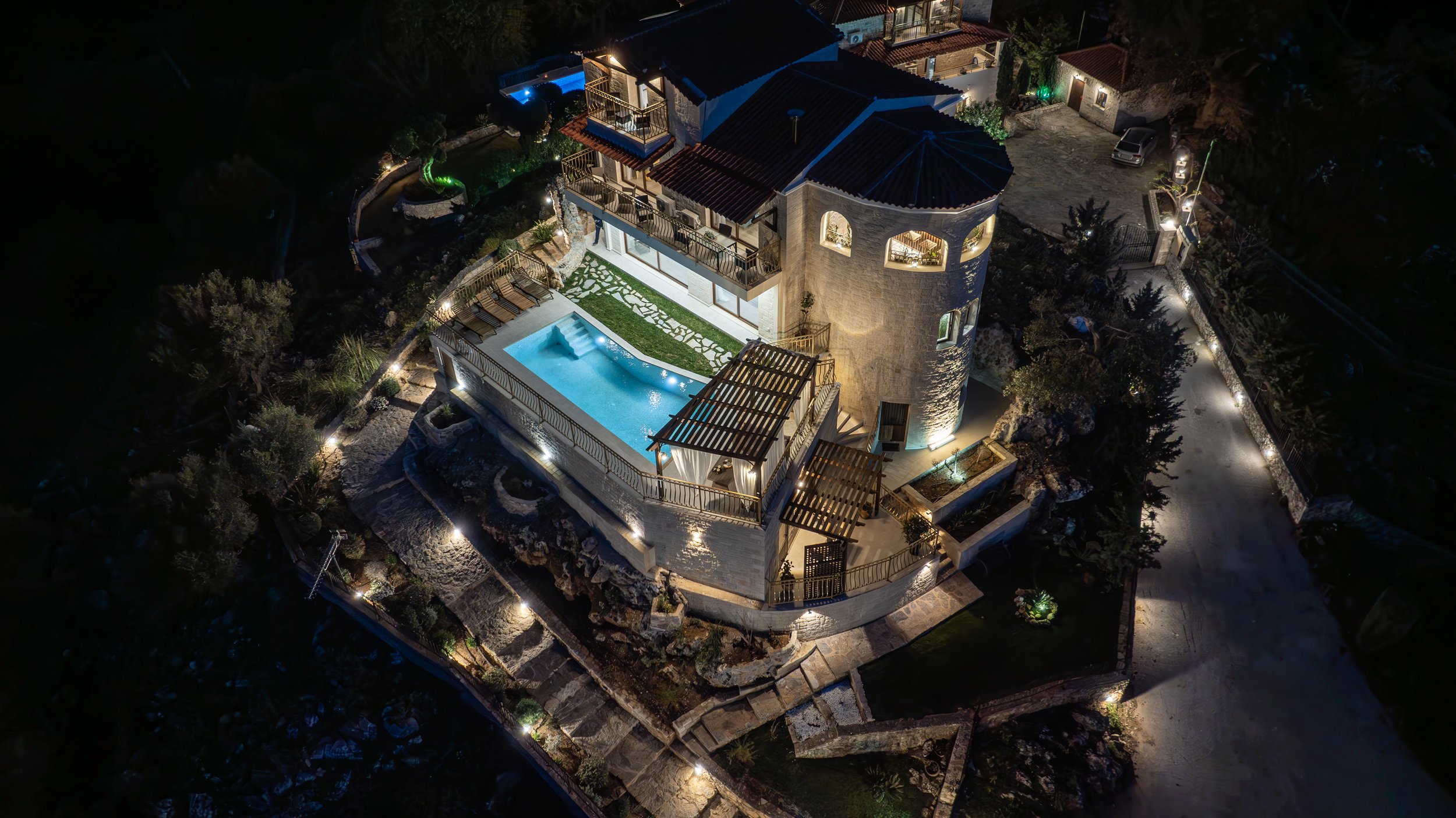 Aerial night view of a luxurious mansion with a pool, illuminated pathways, and outdoor areas.