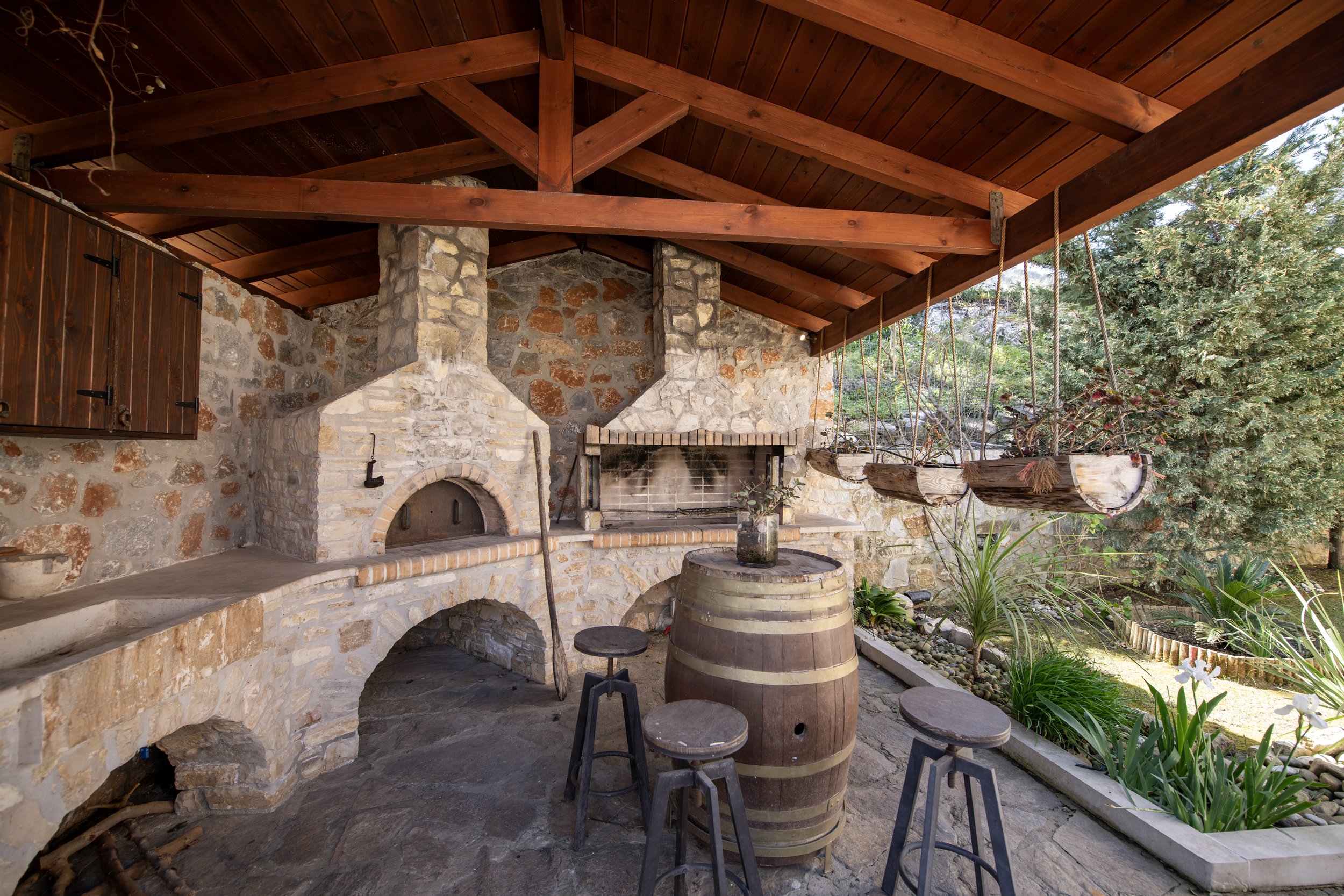 Stone outdoor kitchen with pizza oven and barbecue grill under wooden roof, featuring a barrel table, stools, and hanging planters, set beside a garden area.
